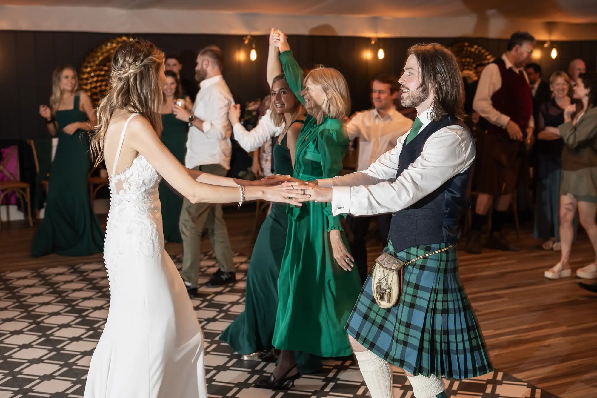 A bride and a man in a kilt dance together at a wedding reception, surrounded by guests dressed in green attire.