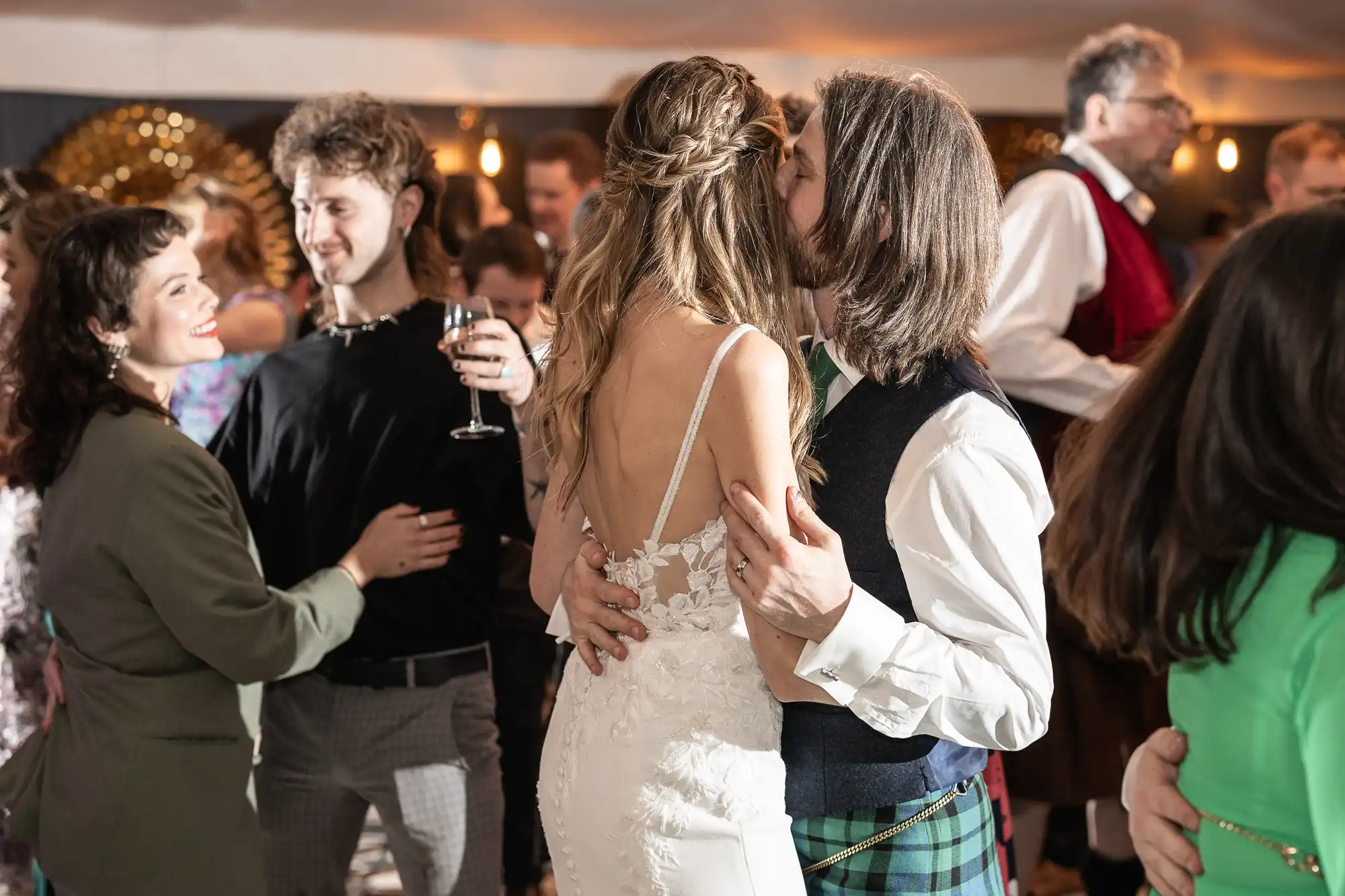 A couple, with the woman in a white dress and the man in a plaid kilt, dance closely at a lively event with other pairs dancing and mingling in the background.