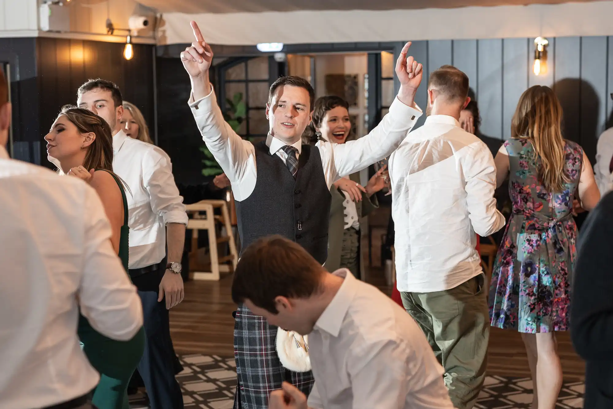 A group of people are dancing at a party. A man in the center wears a vest and plaid pants, raising both hands with fingers pointing up. Others around him are mingling and dancing.