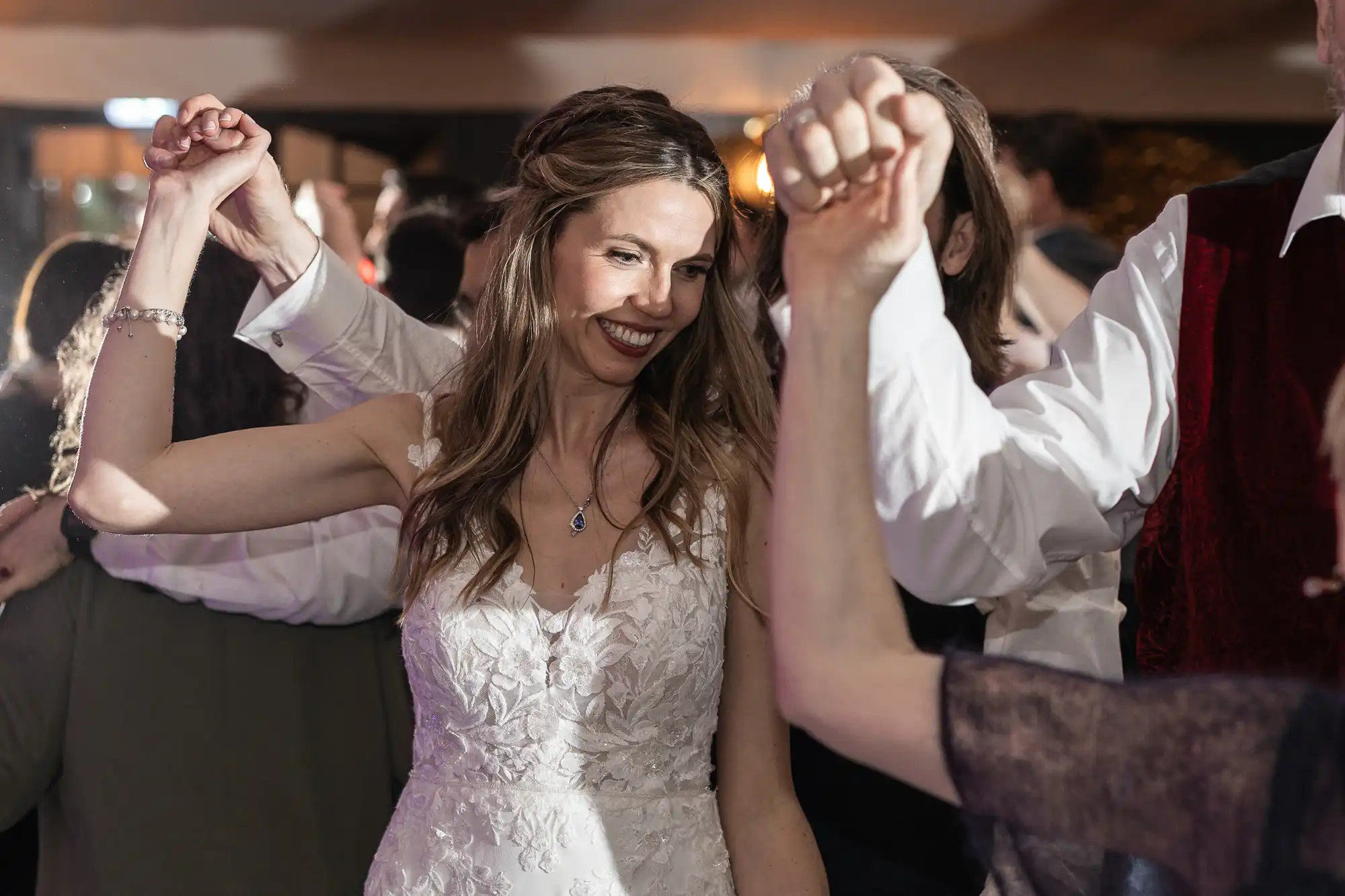 A bride in a white dress smiles while holding hands with others in a dancing circle at a wedding reception.