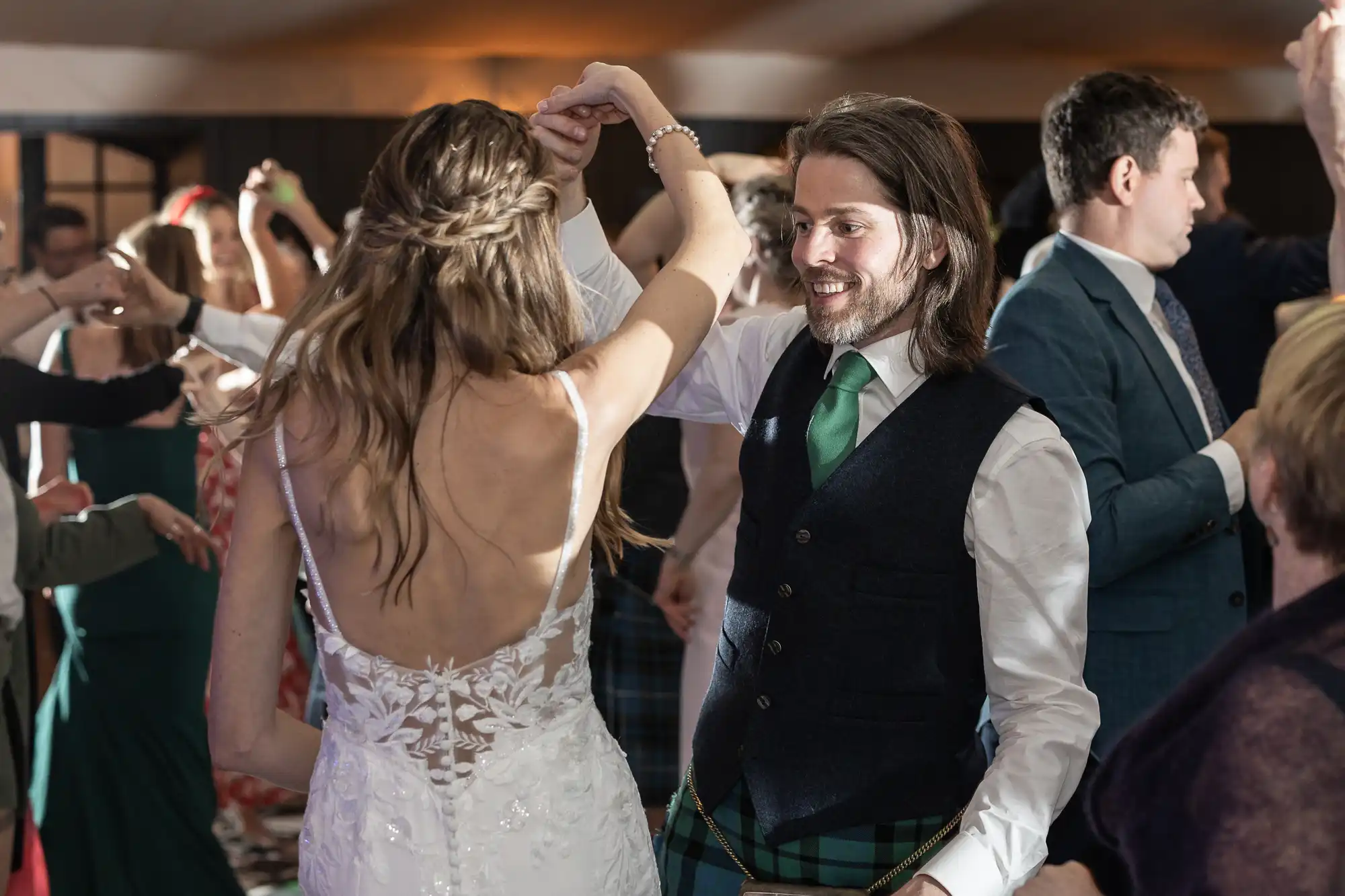 A couple dances, with the woman in a white dress and the man in a suit and green tie, surrounded by others also dancing in a warmly lit room.