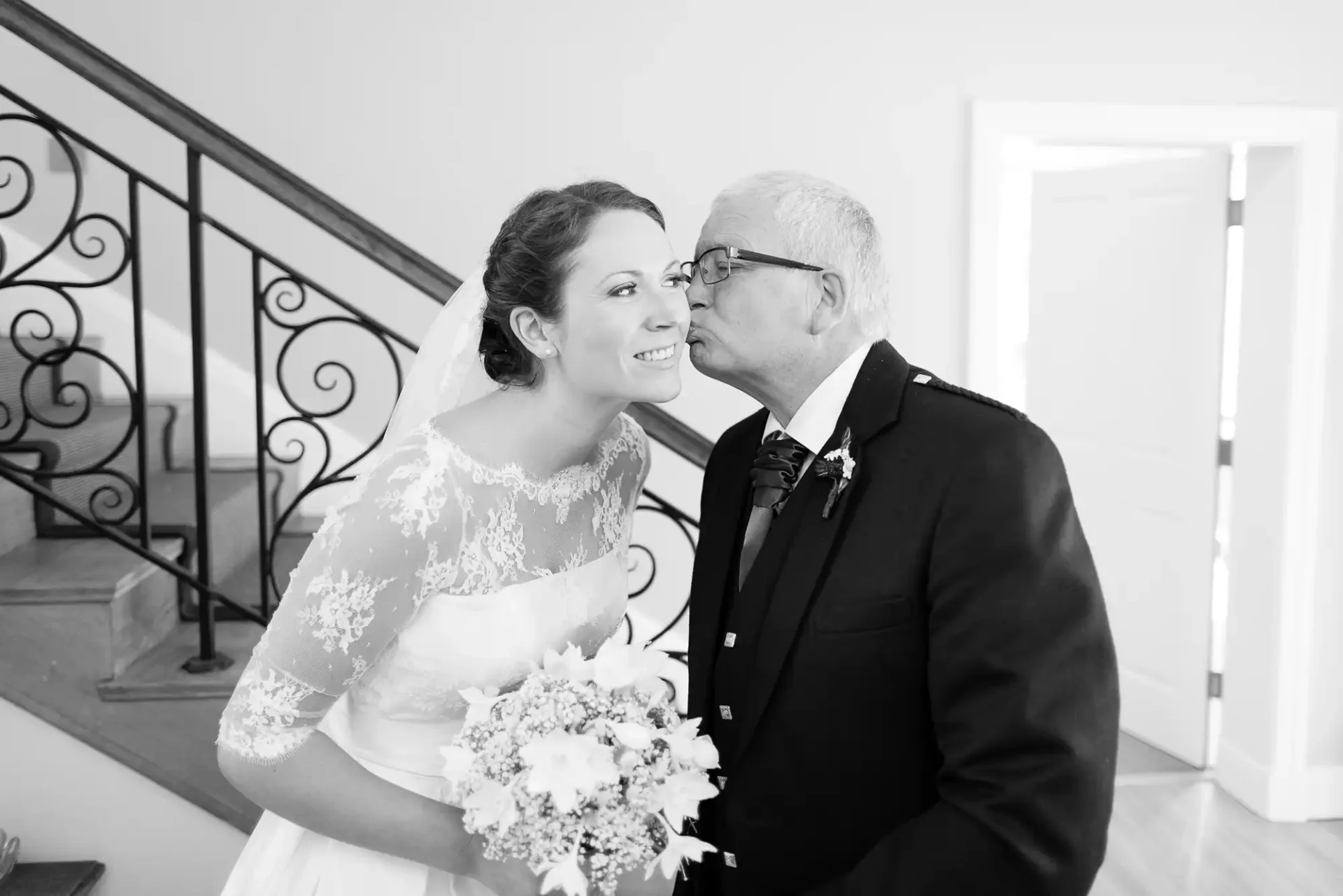 A bride in a lace dress holding a bouquet receives a kiss on the cheek from an older man in a tuxedo on a staircase.