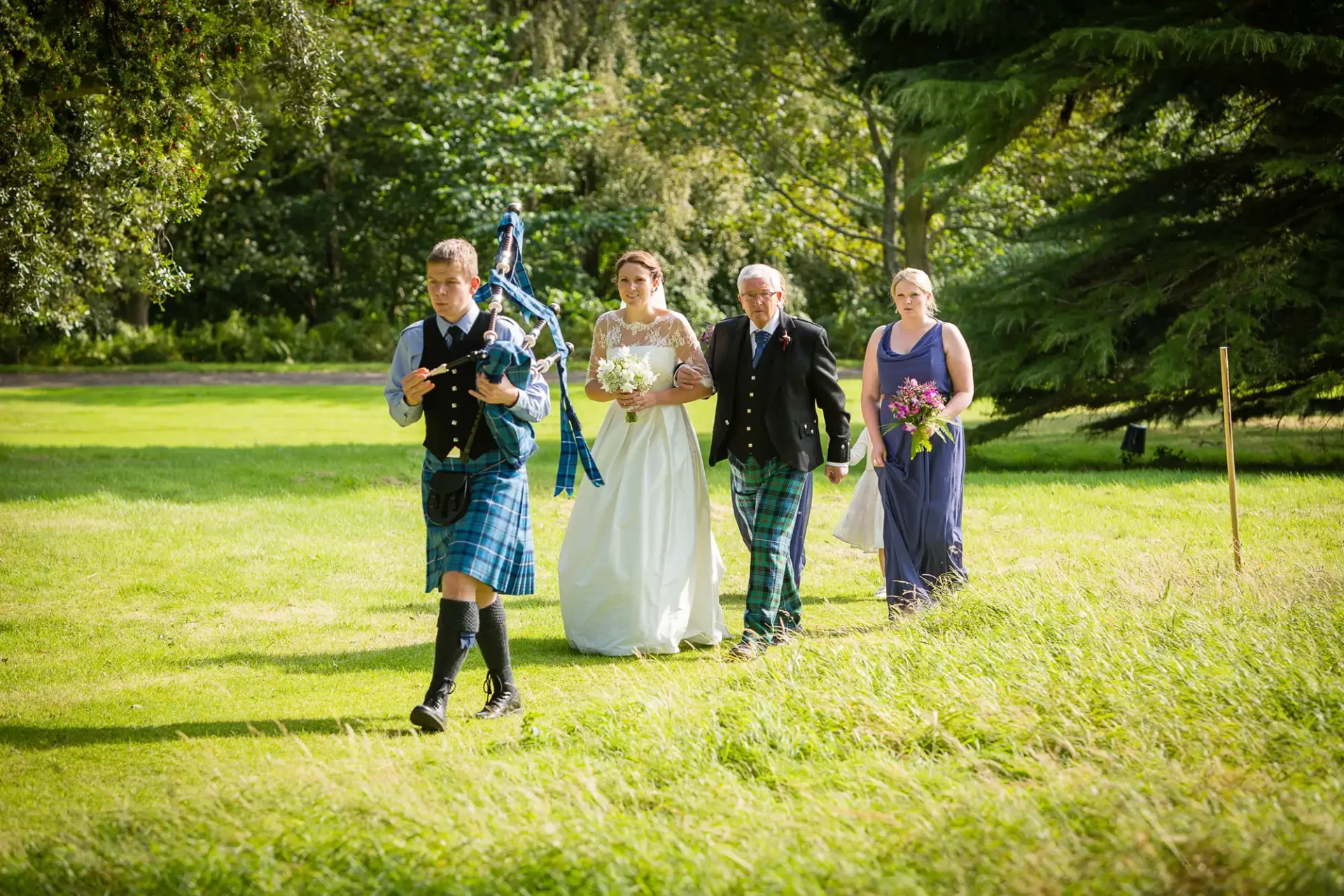 A wedding procession in a park with a bagpiper in a kilt leading a bride and groom, followed by a bridesmaid.