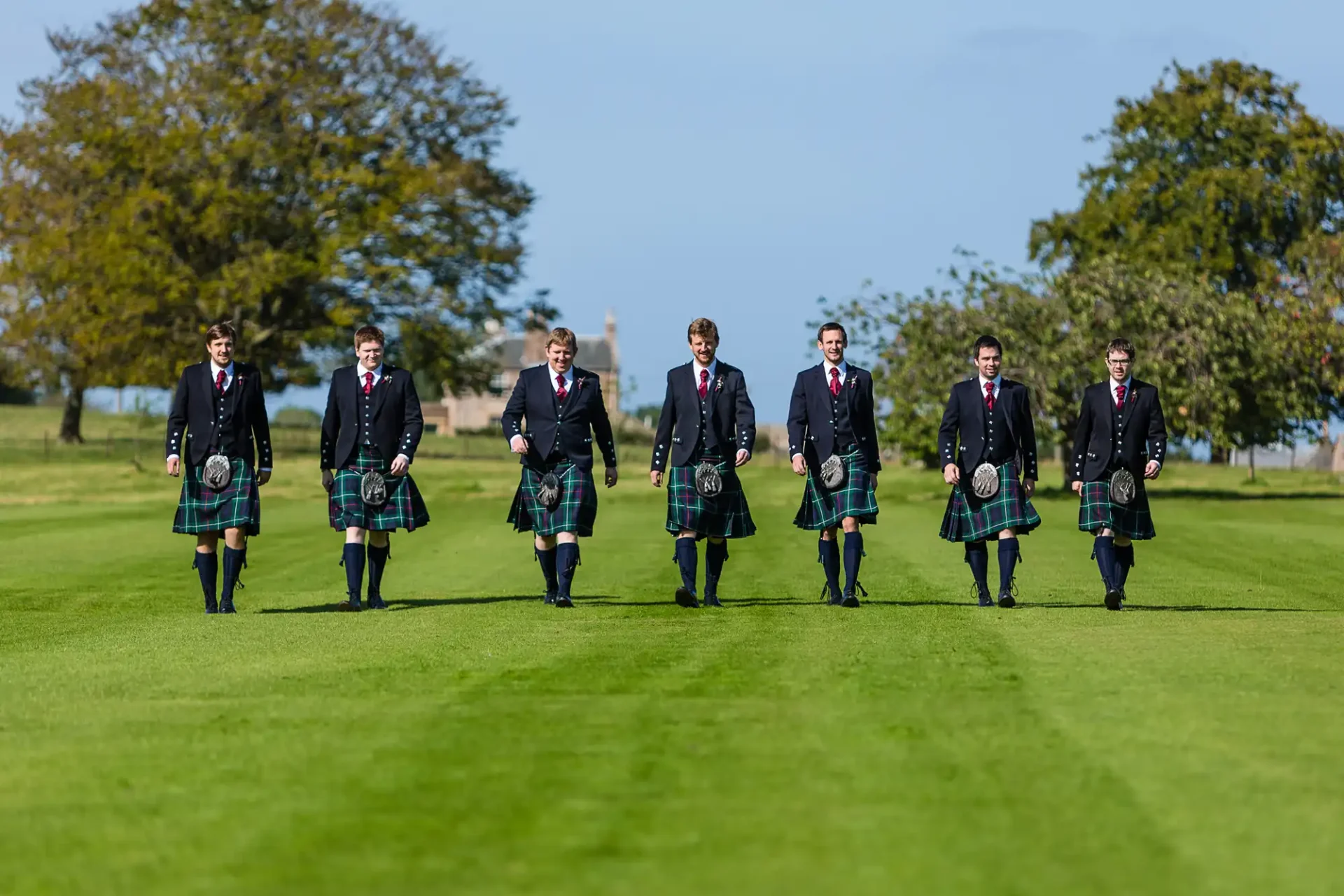 Seven men in traditional scottish kilts and jackets walking in a line on a lush green lawn.