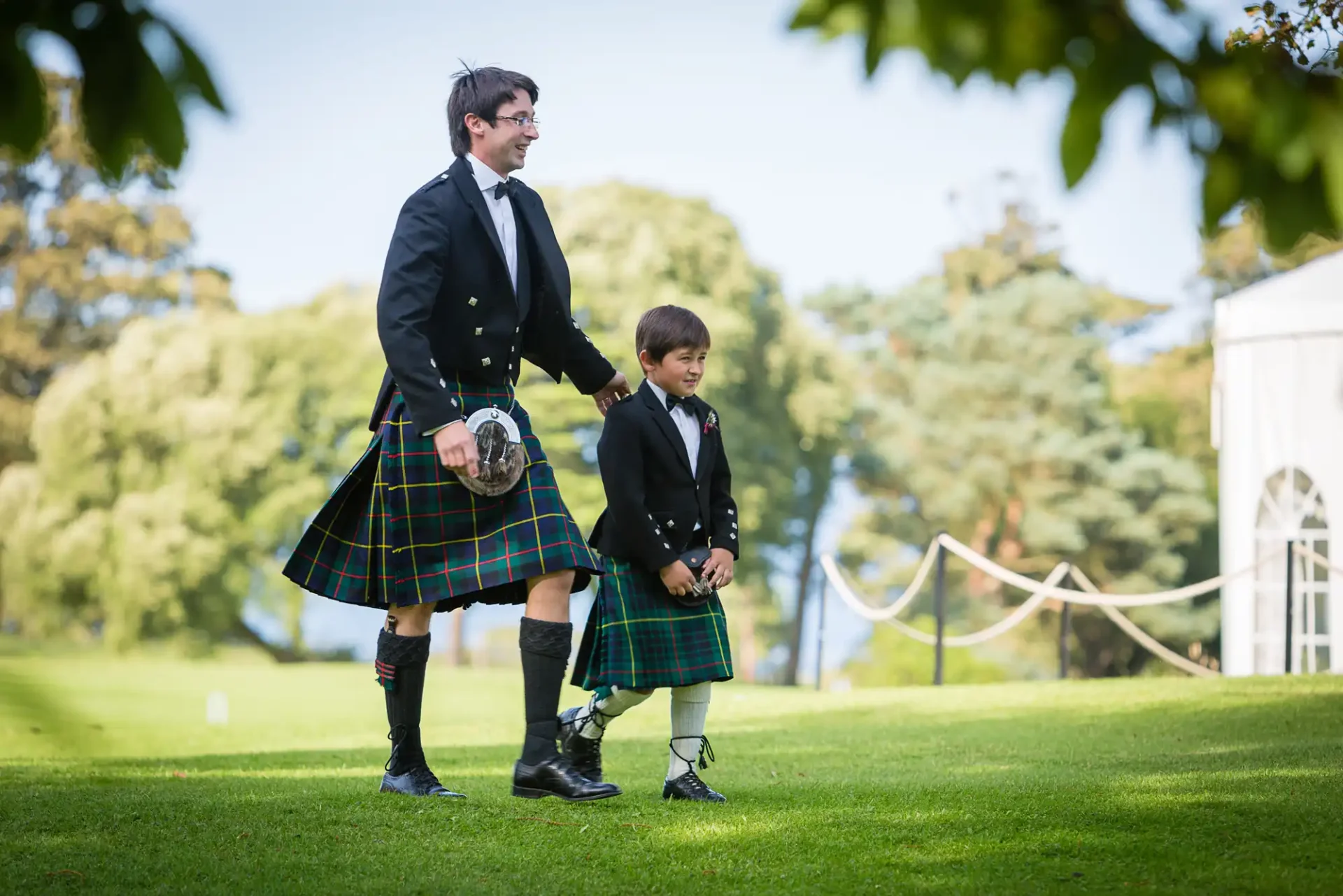 A man and a boy, both wearing traditional tartan kilts and black jackets, walk hand in hand in a sunny park.