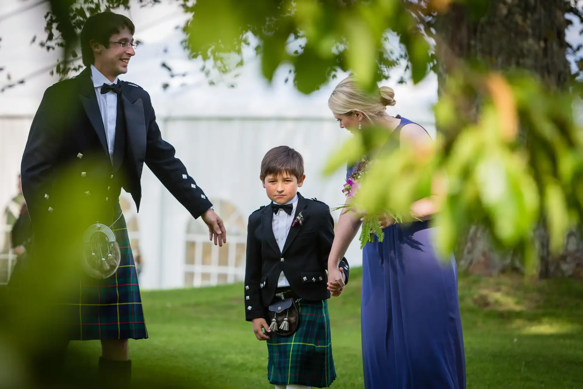 A man and a woman in formal wear interact with a young boy in a kilt at an outdoor event, with bubbles visible around them.