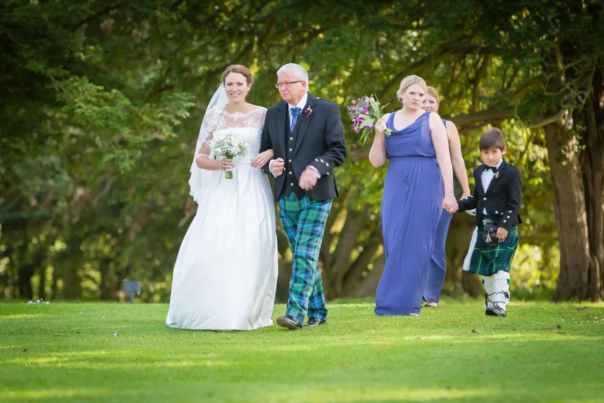 A bride in a white gown, a man in a tartan kilt, a woman in a purple dress, and a young boy in a kilt walk on a grassy path, smiling.