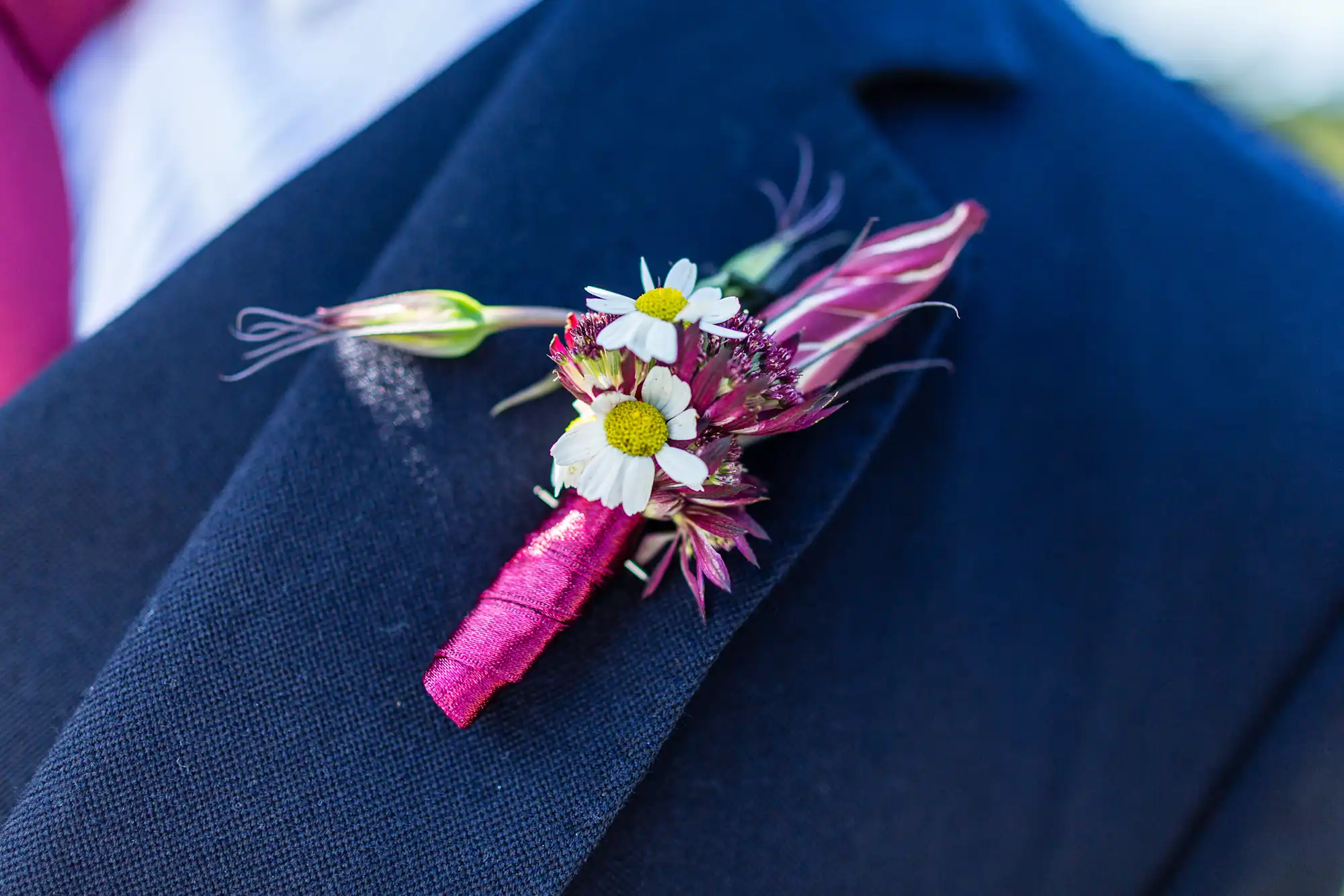 A close-up of a boutonniere with daisies and pink accents on a navy blue suit jacket.