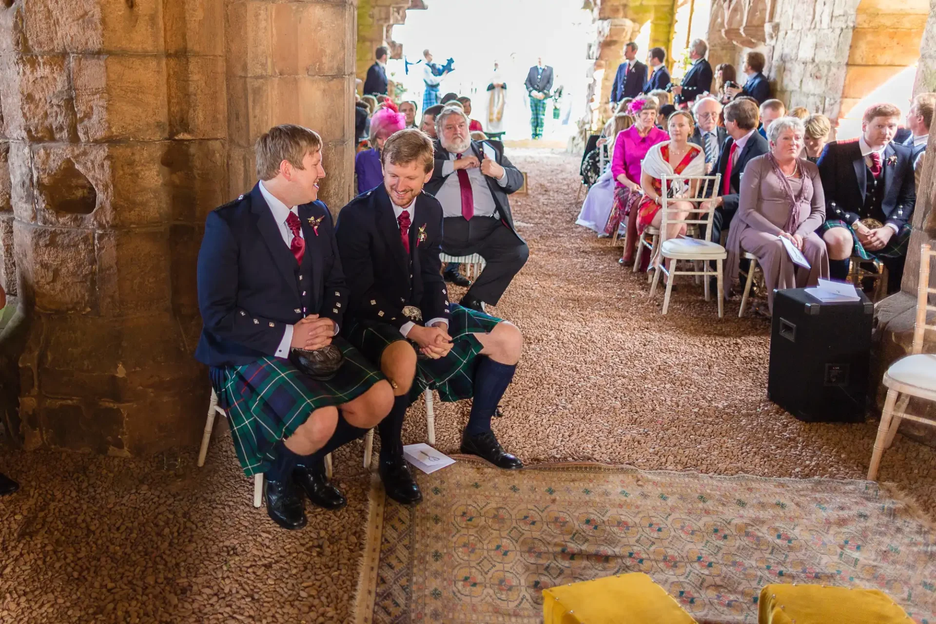 Two men in kilts sitting and chatting inside a historic building with guests in the background at a wedding ceremony.