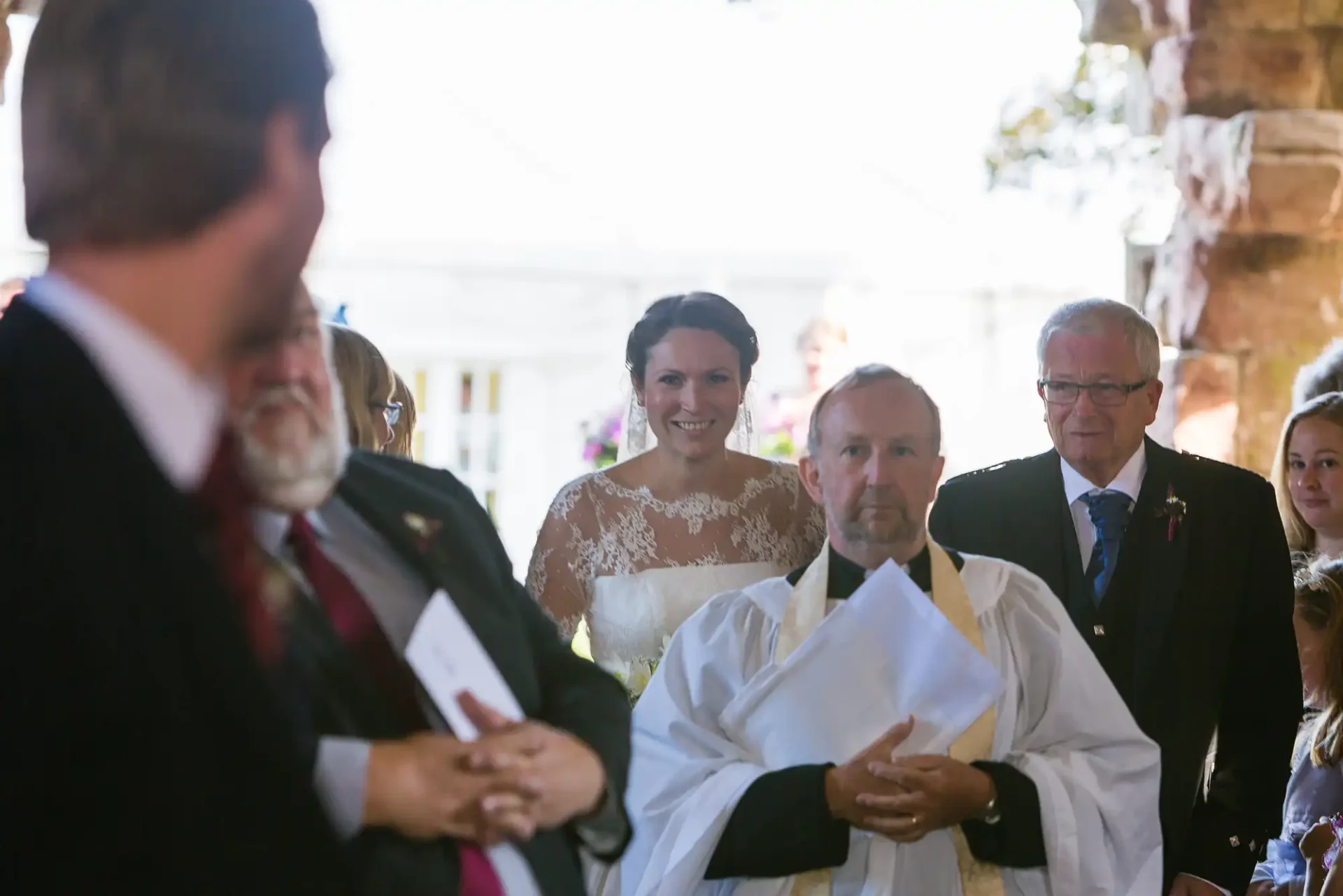A bride walks down the aisle, smiling, accompanied by an older man; a priest and guests are in the foreground.