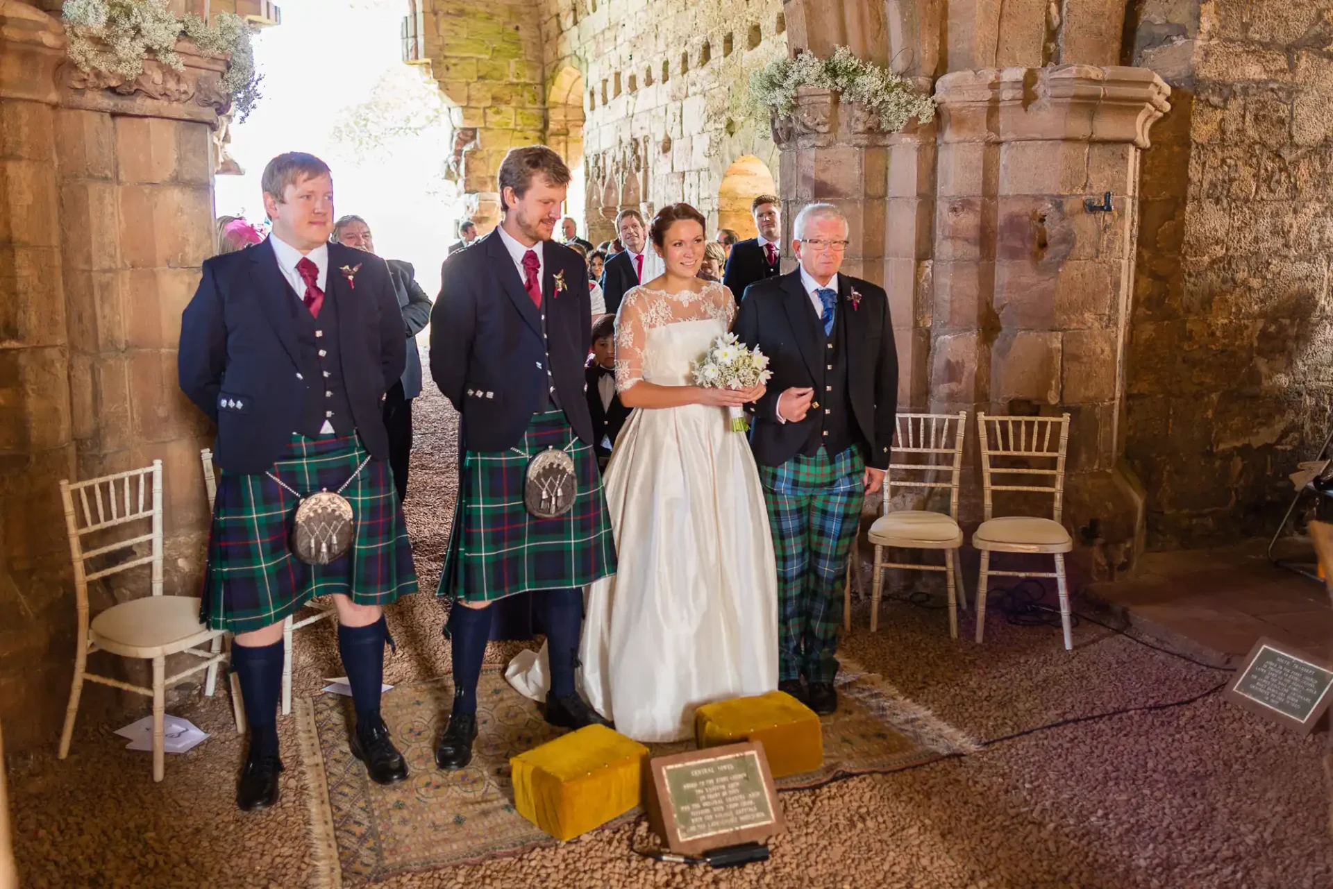 A wedding ceremony inside a historic stone building with the bride and groom walking down the aisle, accompanied by men in traditional kilts.