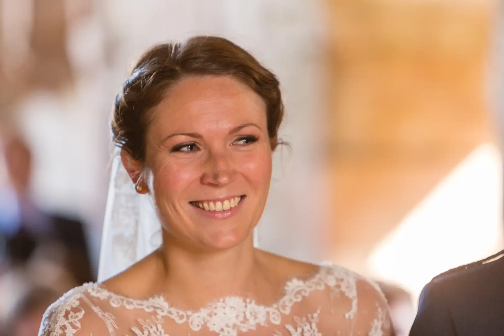 A smiling bride wearing a lace wedding dress and veil, with soft focus and warm lighting in the background.
