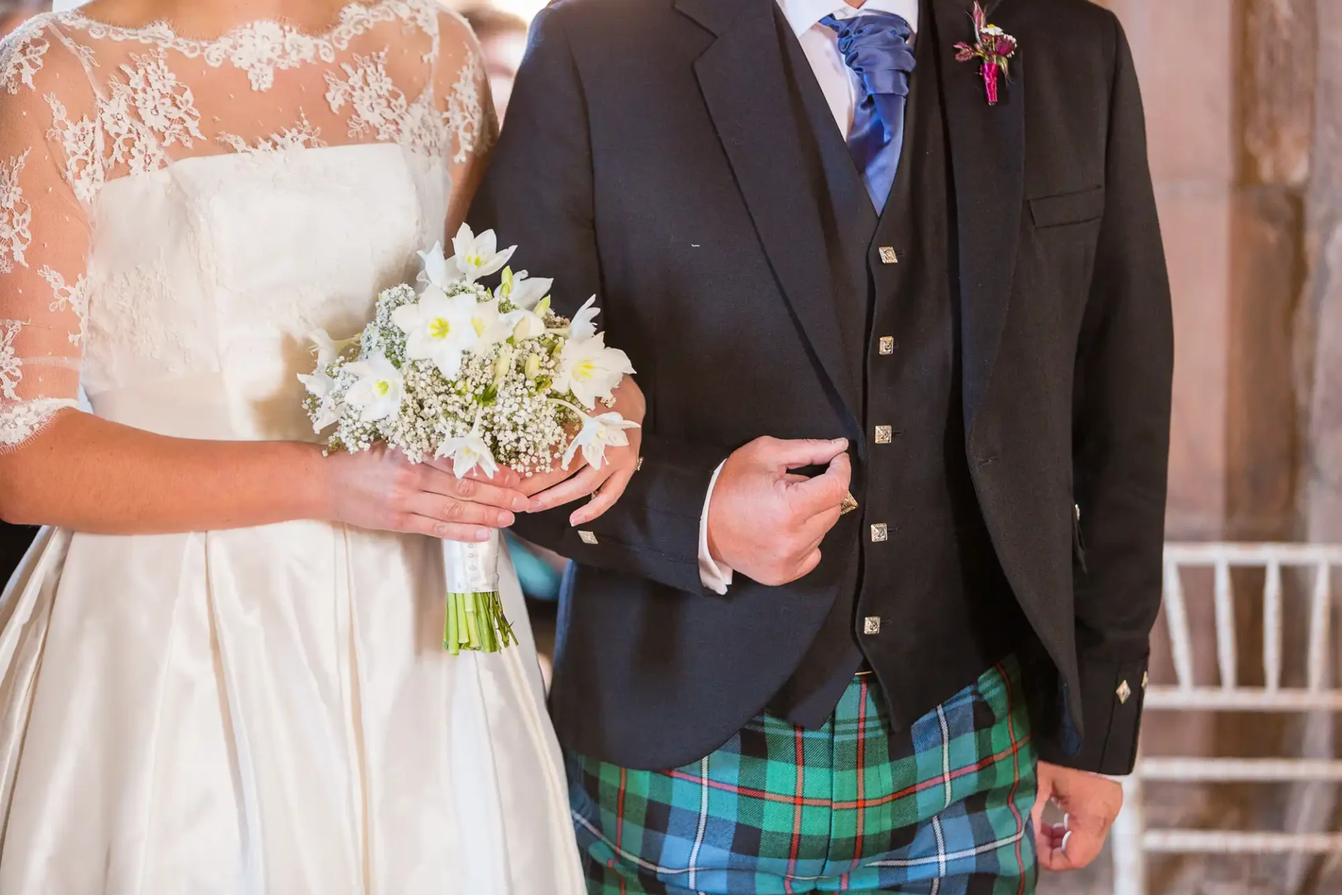 Bride in a lace dress and groom in a tartan kilt holding hands and a white bouquet at a wedding ceremony.
