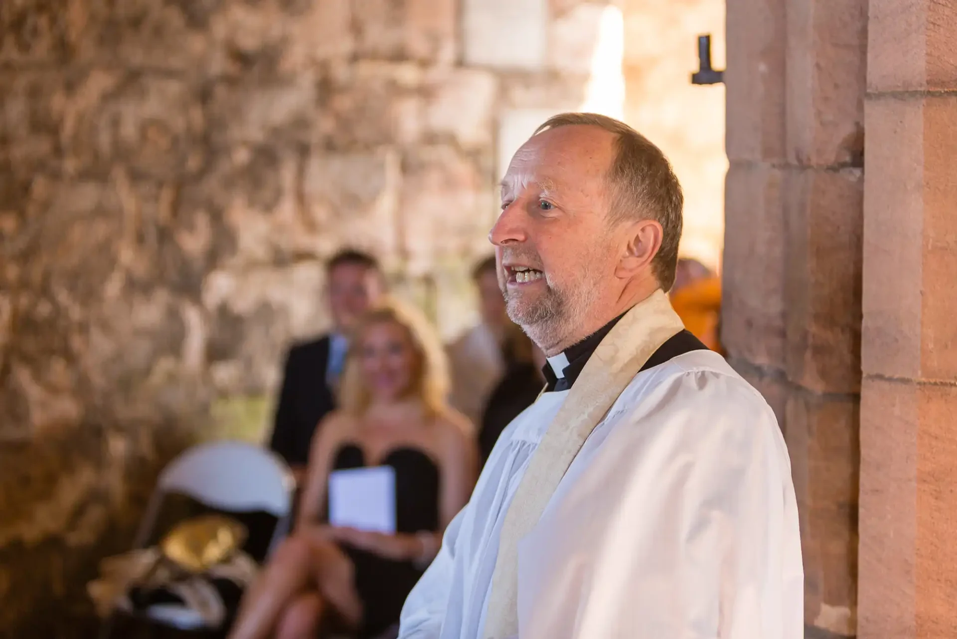 A priest in white vestments speaking at a wedding ceremony in a rustic stone building, with guests visible in the background.