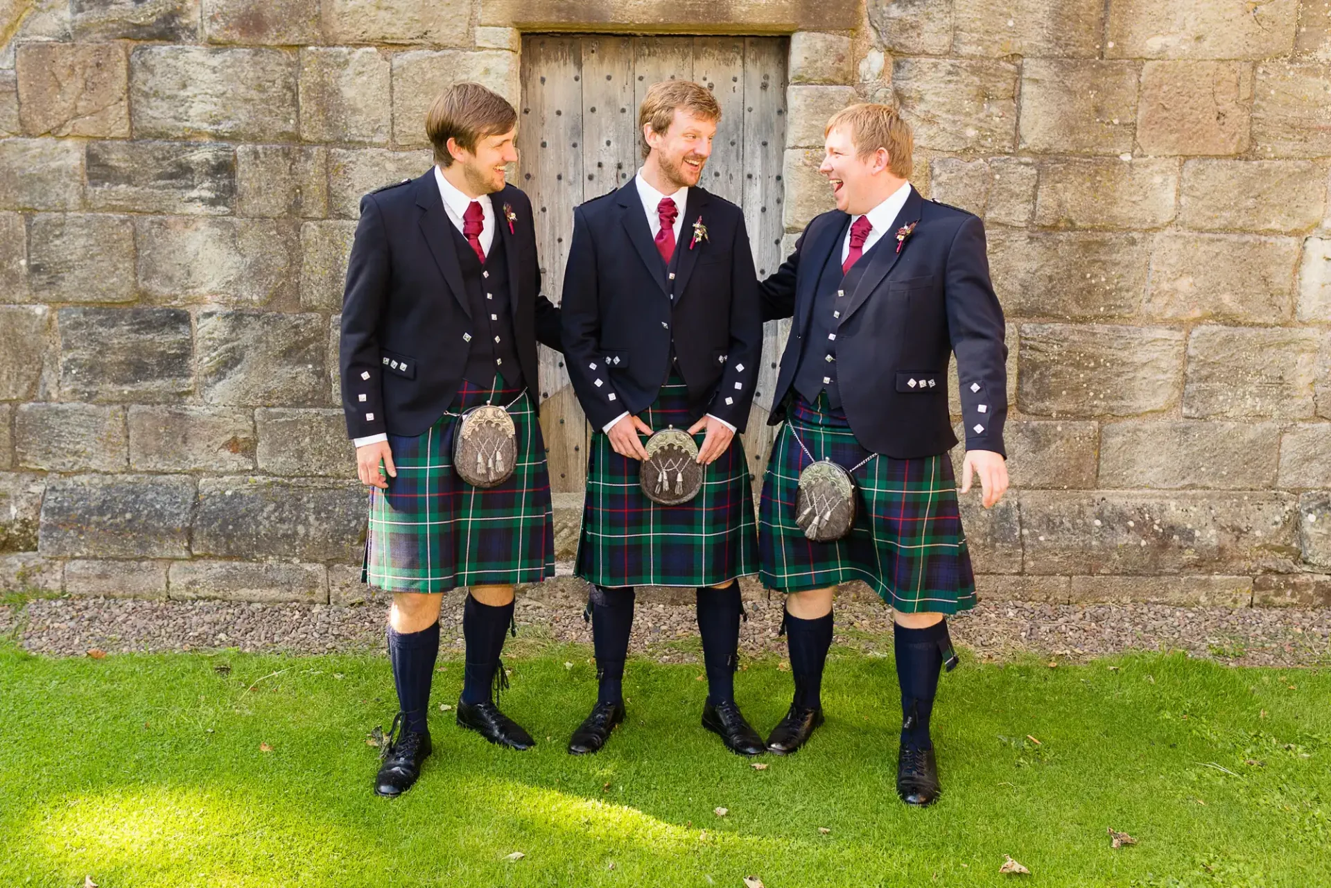 Three men in traditional scottish kilts and jackets, smiling and conversing outside a stone building.
