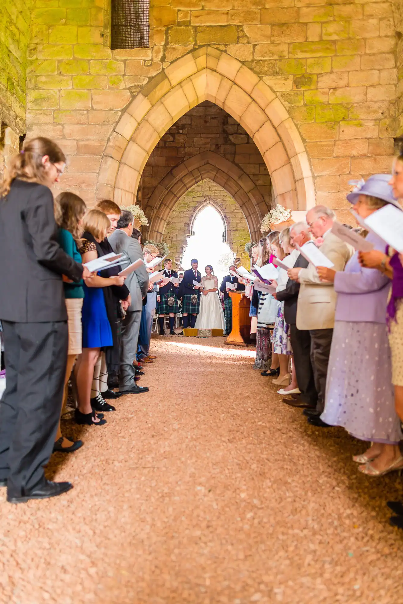 A wedding ceremony in a medieval stone archway, with guests lined up on each side holding hymn sheets.