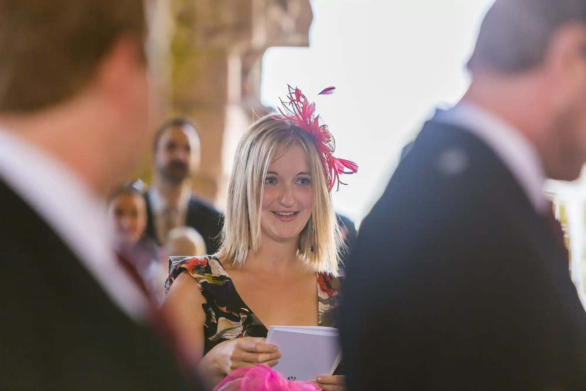 A woman in a floral dress and red fascinator smiles while holding a program at a social event, surrounded by other guests.