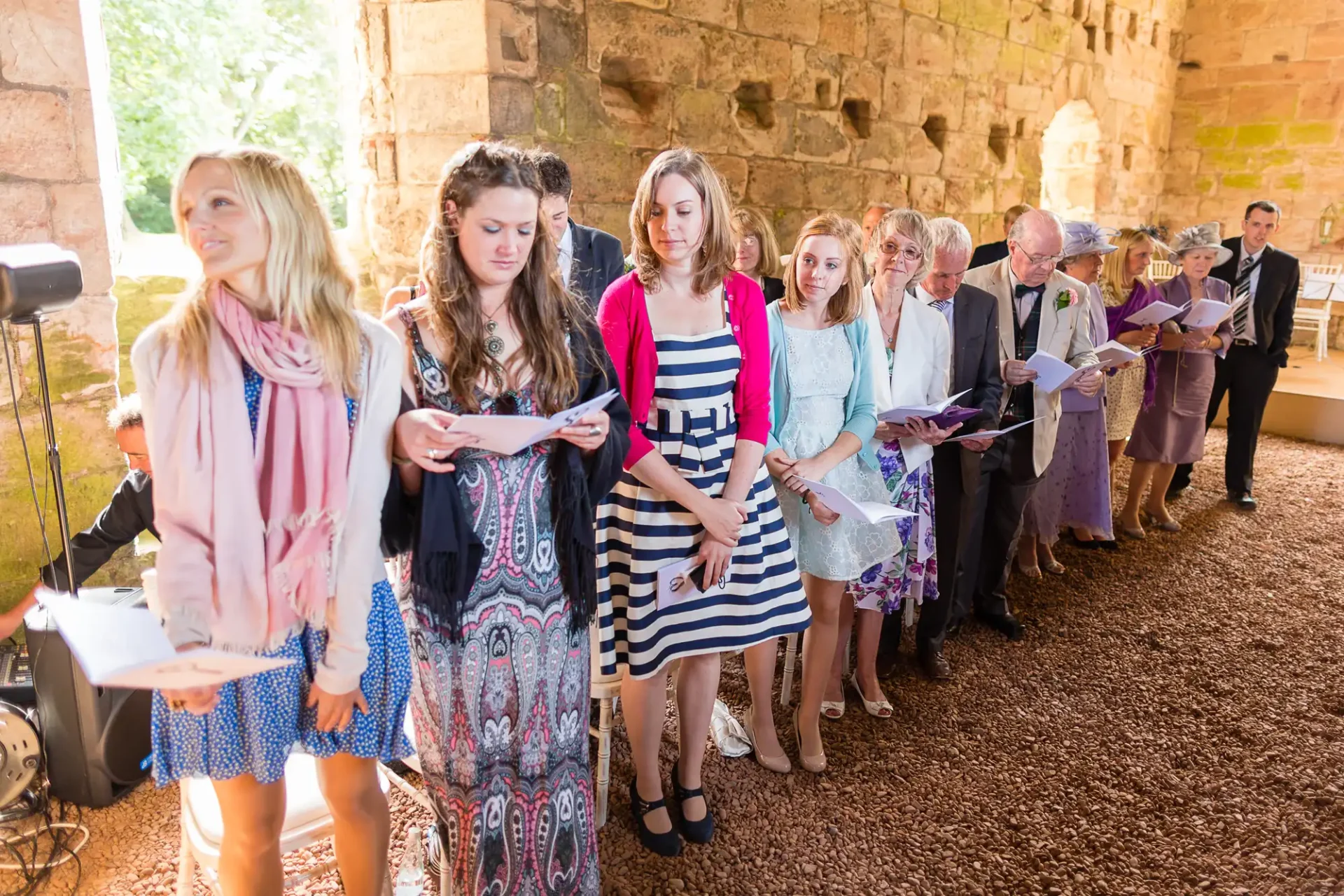 Group of people standing and singing from hymn sheets at a ceremony inside a stone-walled building with sunlight streaming through.