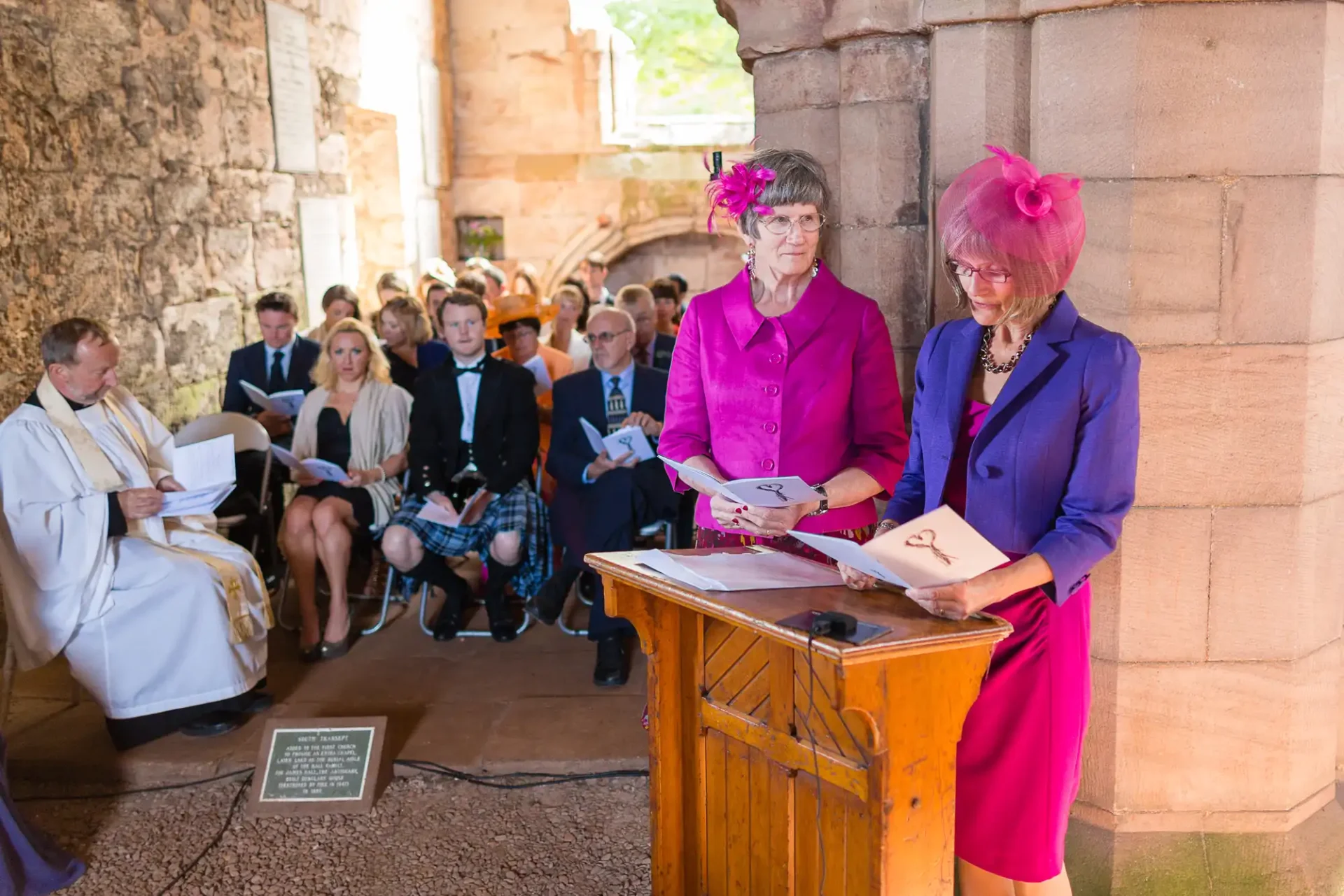 Two women in vibrant purple outfits are reading from papers at a lectern during a ceremony, with guests seated in the background.