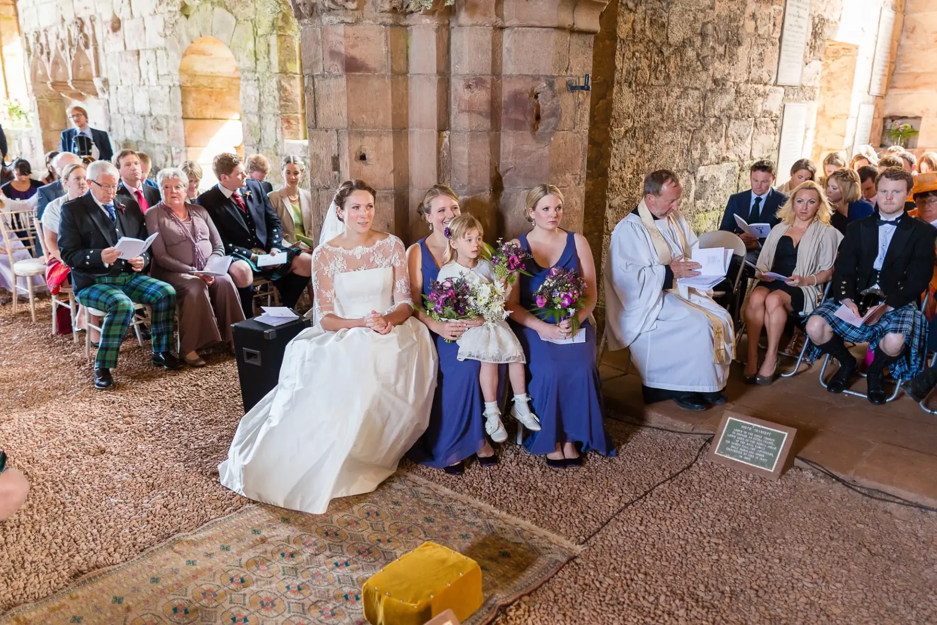 A wedding ceremony in a historic venue with guests in traditional and formal attire, featuring a bride in white and attendees in various colorful outfits.