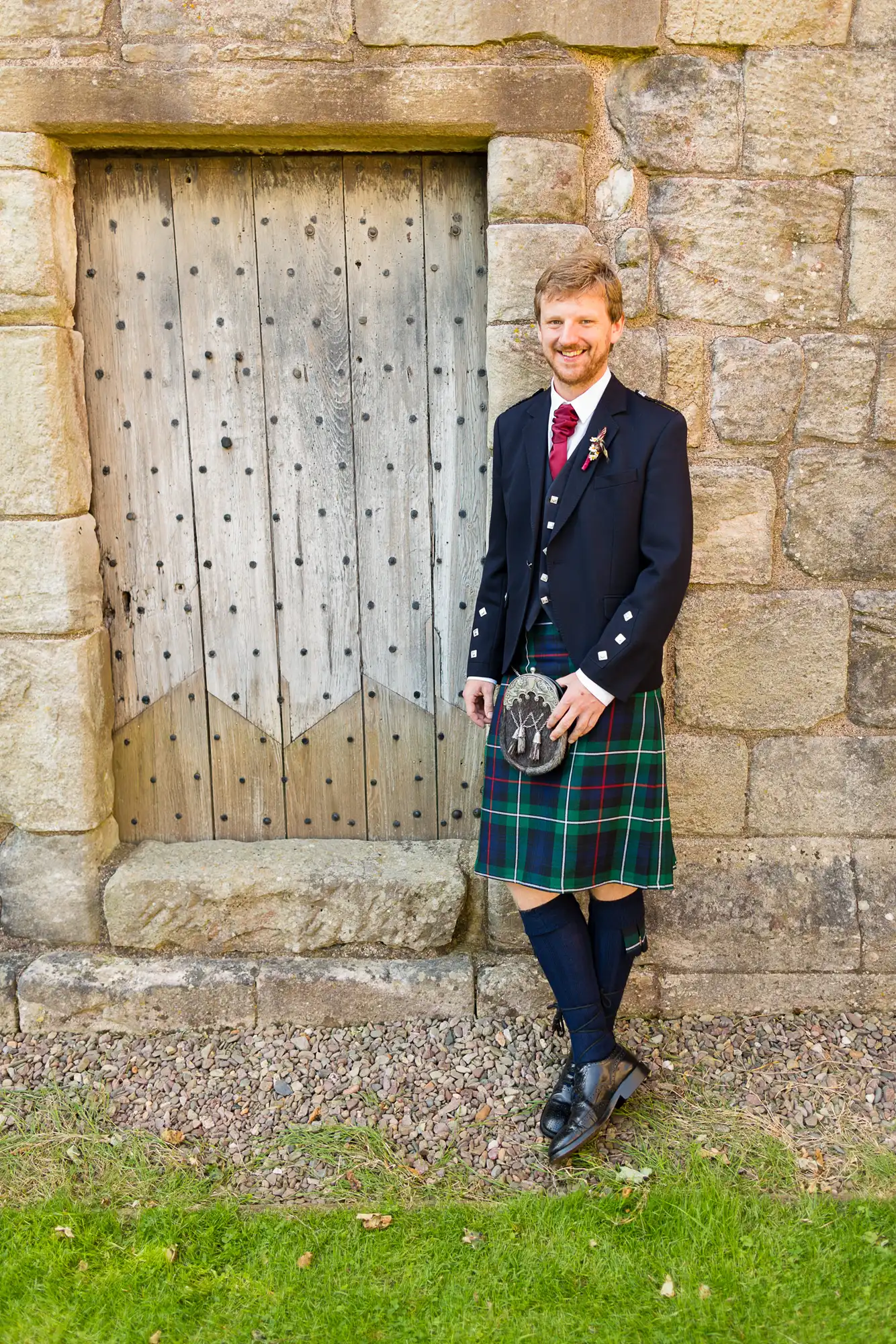Man in traditional scottish attire, including kilt and sporran, standing beside an old wooden door, smiling.