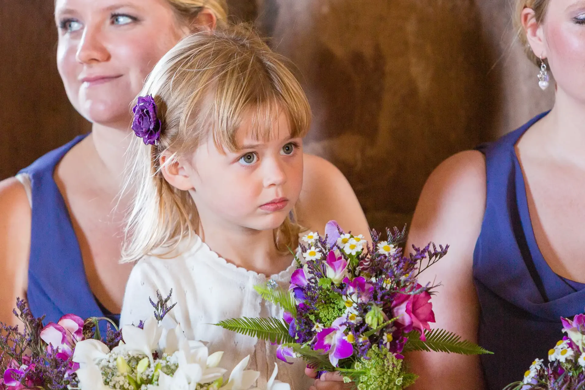 A young girl with a white dress and a purple flower in her hair holds a bouquet, sitting beside two women in blue dresses at a formal event.