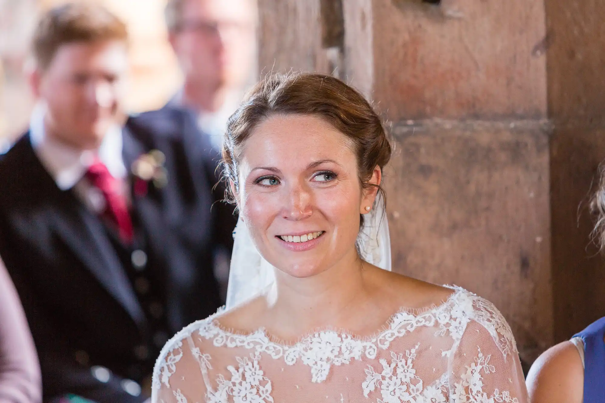 A bride in a lace wedding dress smiling softly during a ceremony, with out-of-focus guests in the background.
