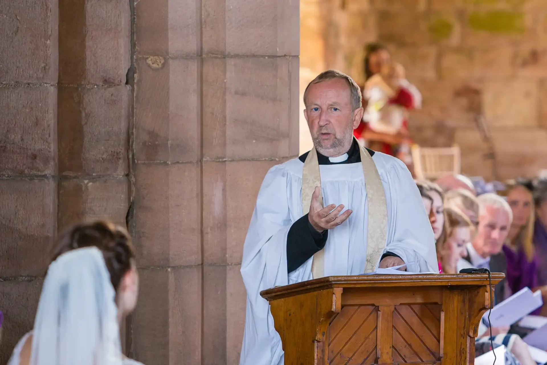 A priest in white robes delivering a sermon from a wooden pulpit in a church, with listeners in the background.