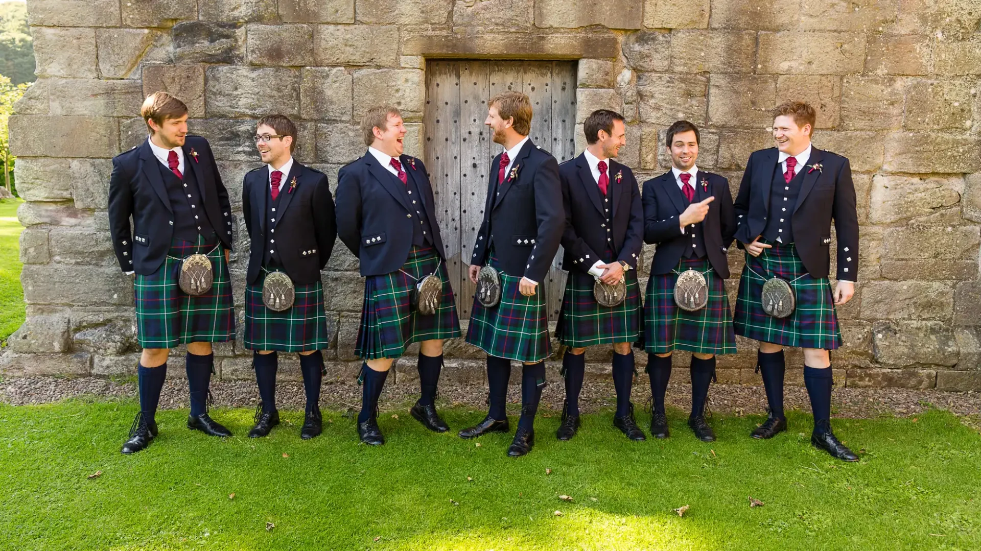 Group of seven men in matching dark blazers and tartan kilts, standing and laughing in front of an old stone building.