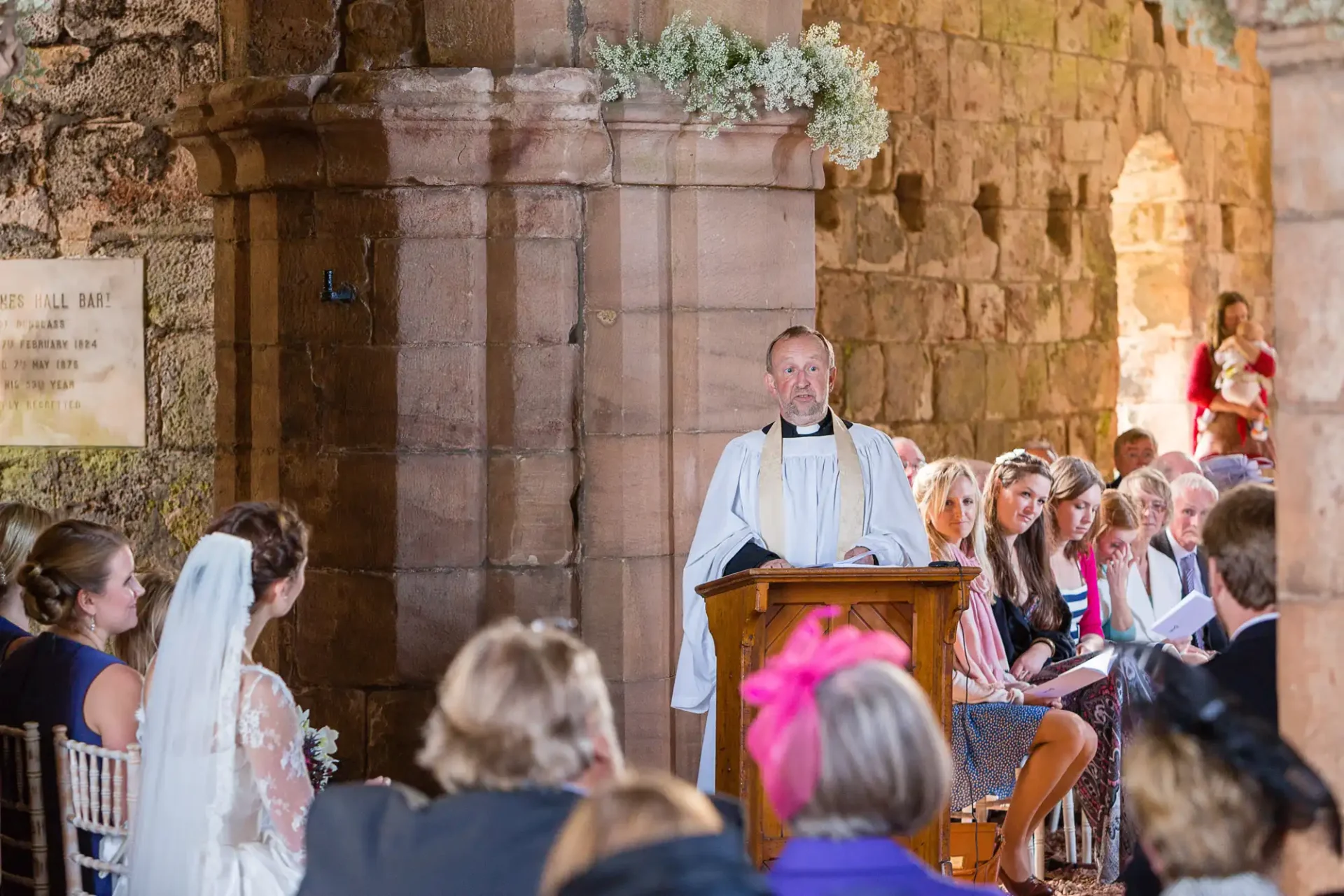 Priest speaking at a lectern during a wedding ceremony in a historic stone church, with guests seated and a bride listening attentively.