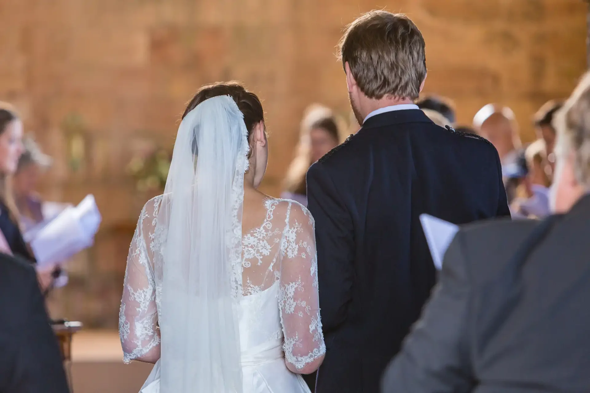 Back view of a bride in a lace gown and a groom in a suit, standing together during their wedding ceremony inside a church.