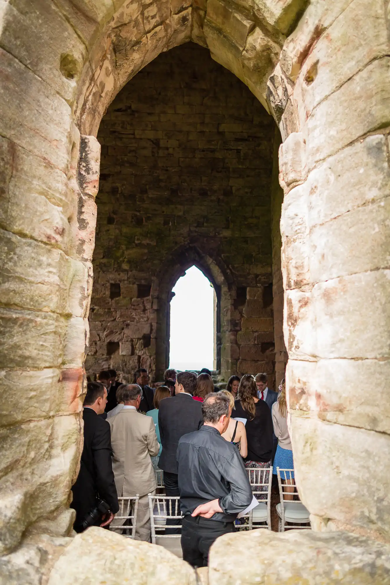 Group of people seated at a wedding ceremony inside an ancient stone building with a large arched window.