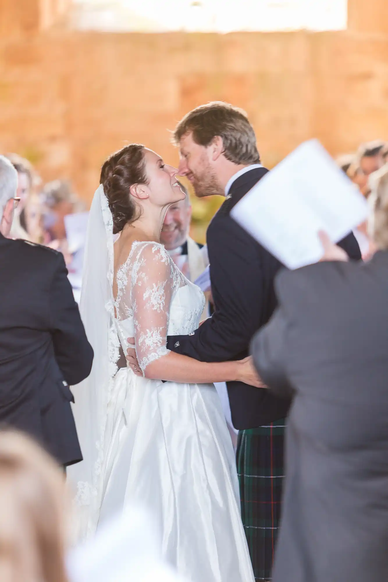A bride and groom share a kiss, the bride in a white lace gown and the groom in a kilt, surrounded by wedding guests in a warmly lit setting.
