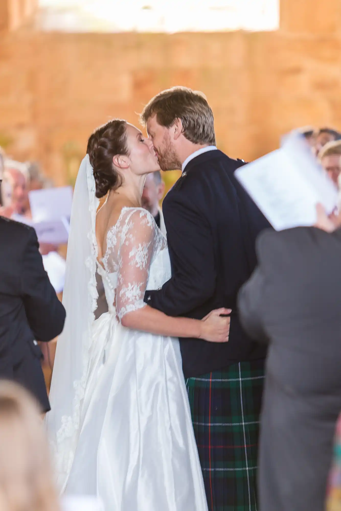 A bride in a lace gown and a groom in a kilt kiss surrounded by guests at a wedding ceremony.