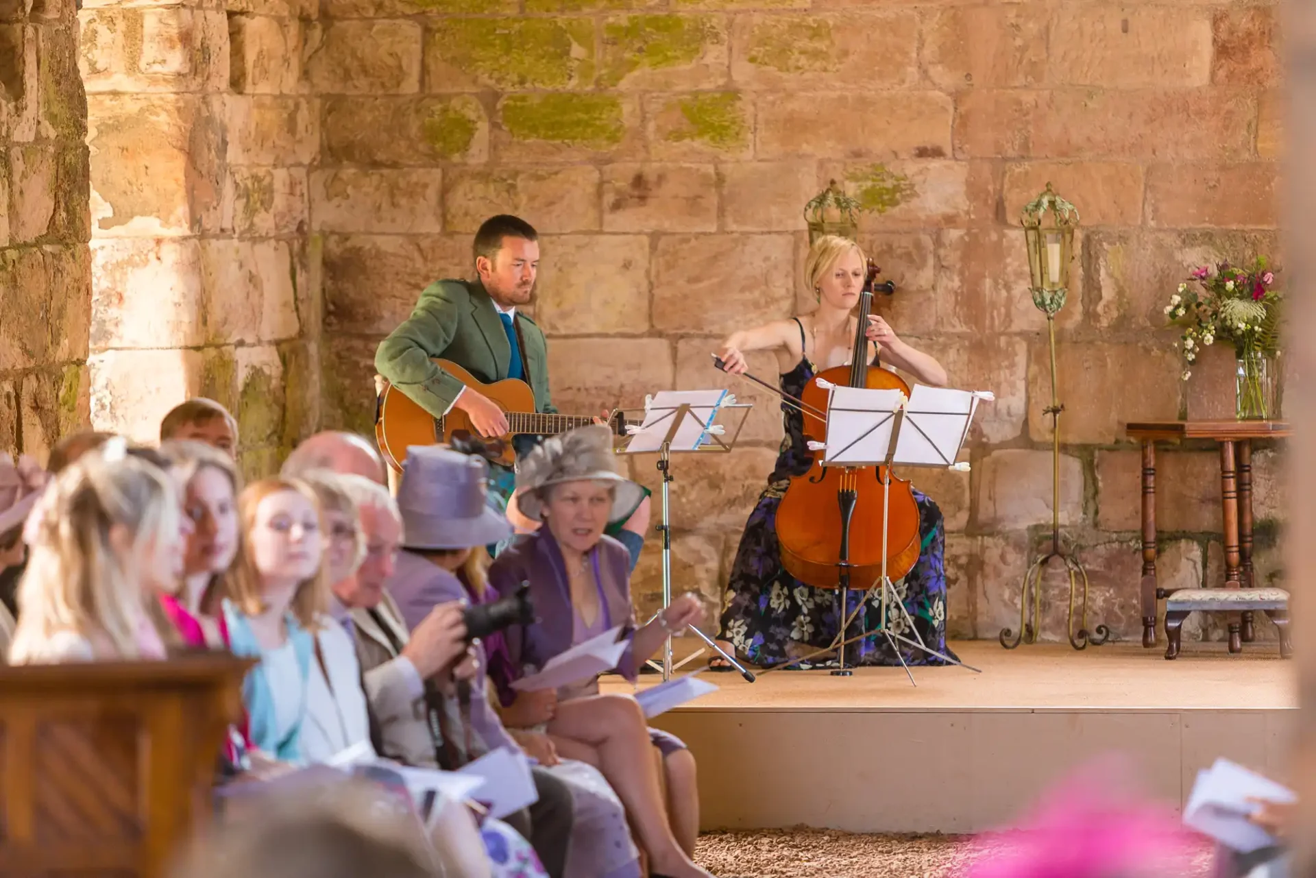 Two musicians, a guitarist and a cellist, perform for an audience at an outdoor event, with listeners seated holding programs.