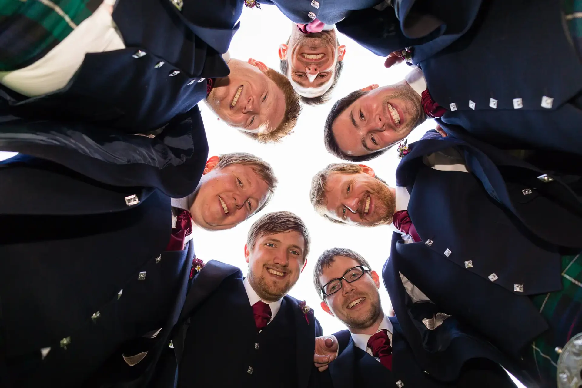 Group of six men in kilts and formal jackets smiling down at the camera, viewed from a low angle.