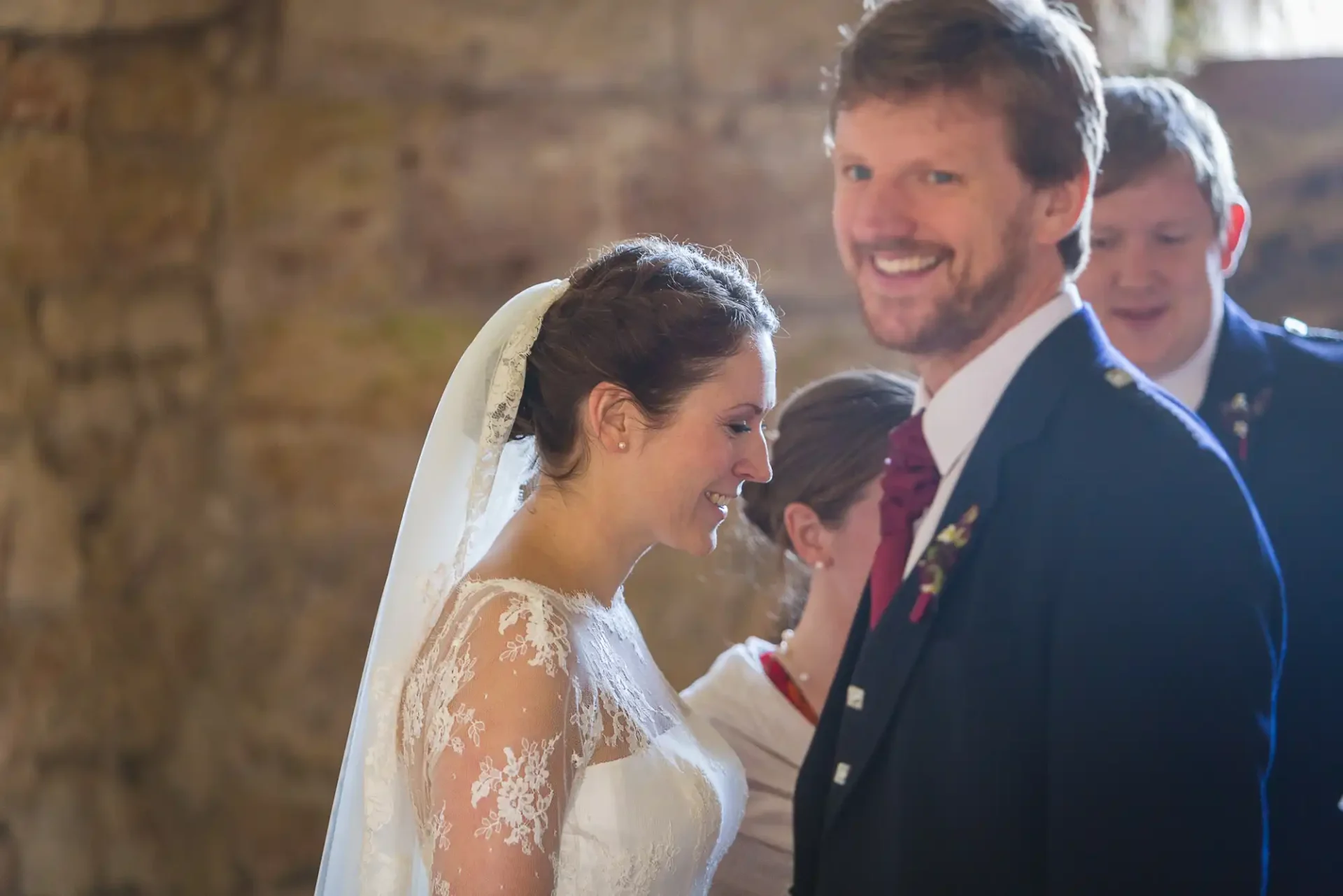 A bride and groom smiling at each other, lit by natural light in an indoor setting, with a guest visible in the background.
