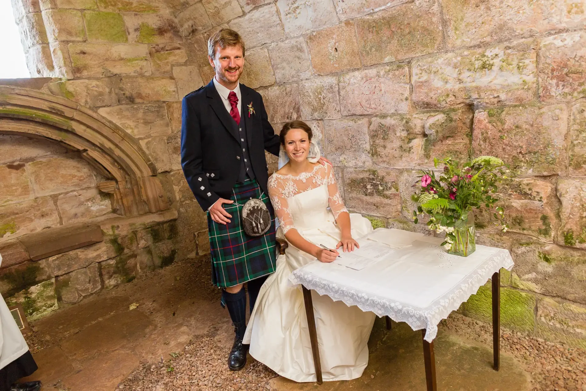 A smiling bride and groom in traditional scottish attire pose beside a stone wall and under an arched window, with a small floral arrangement on a table next to them.