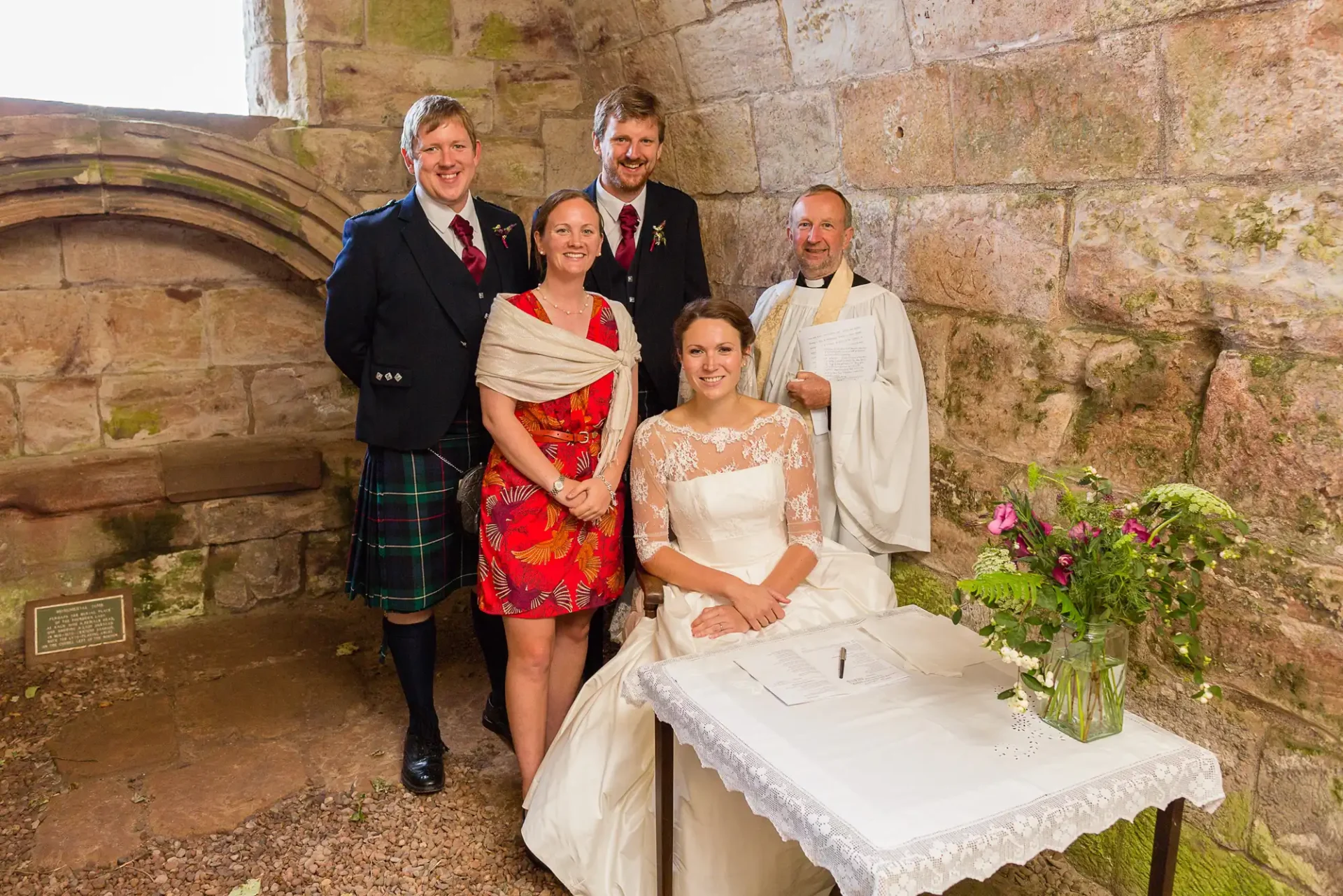 Five people, including a bride and groom, pose with a priest near a signing table adorned with flowers inside a historical stone building.