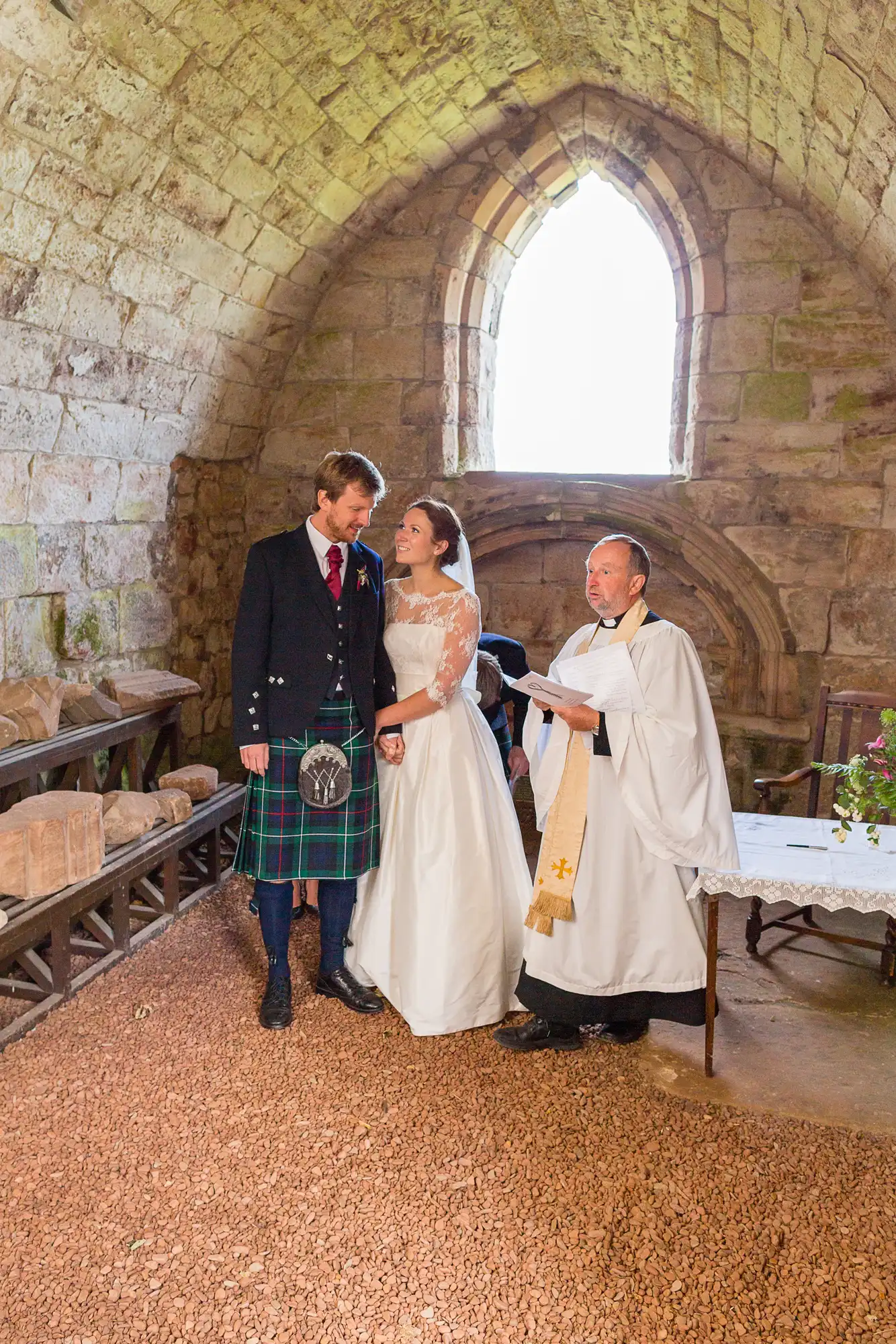 Bride and groom in a stone chapel, with the groom wearing a kilt and the bride in a long white dress, standing beside a priest during their wedding ceremony.