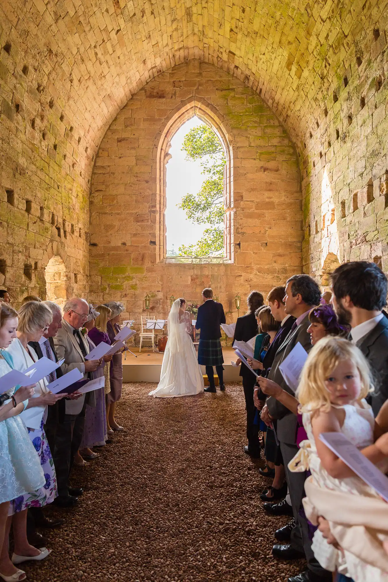 A wedding ceremony inside a historical stone chapel with guests and a couple at the altar, large arched window in the background.