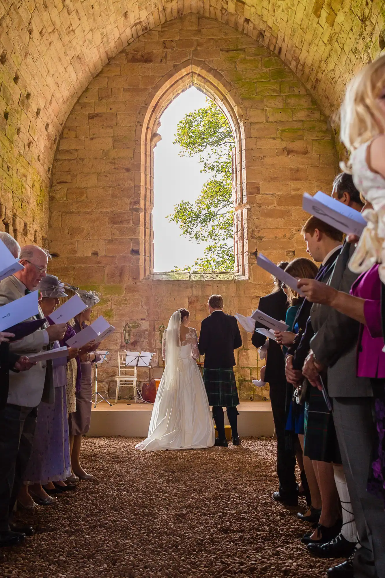 A bride and groom holding hands during a wedding ceremony inside a historic church with a large arched window.