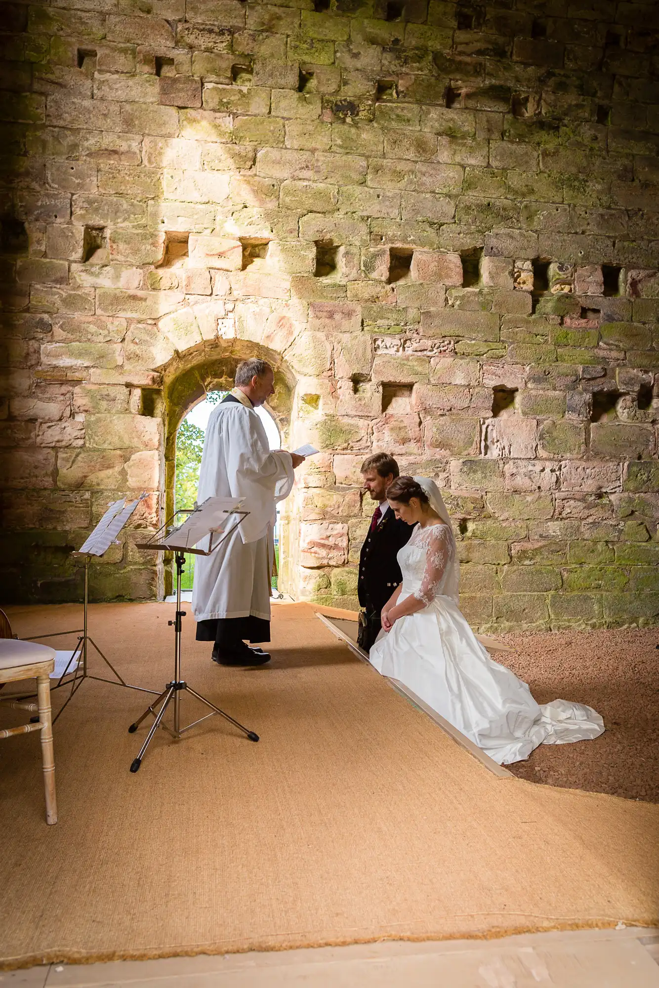 A wedding ceremony in a historic stone building, with a bride and groom kneeling and a priest officiating from a book.