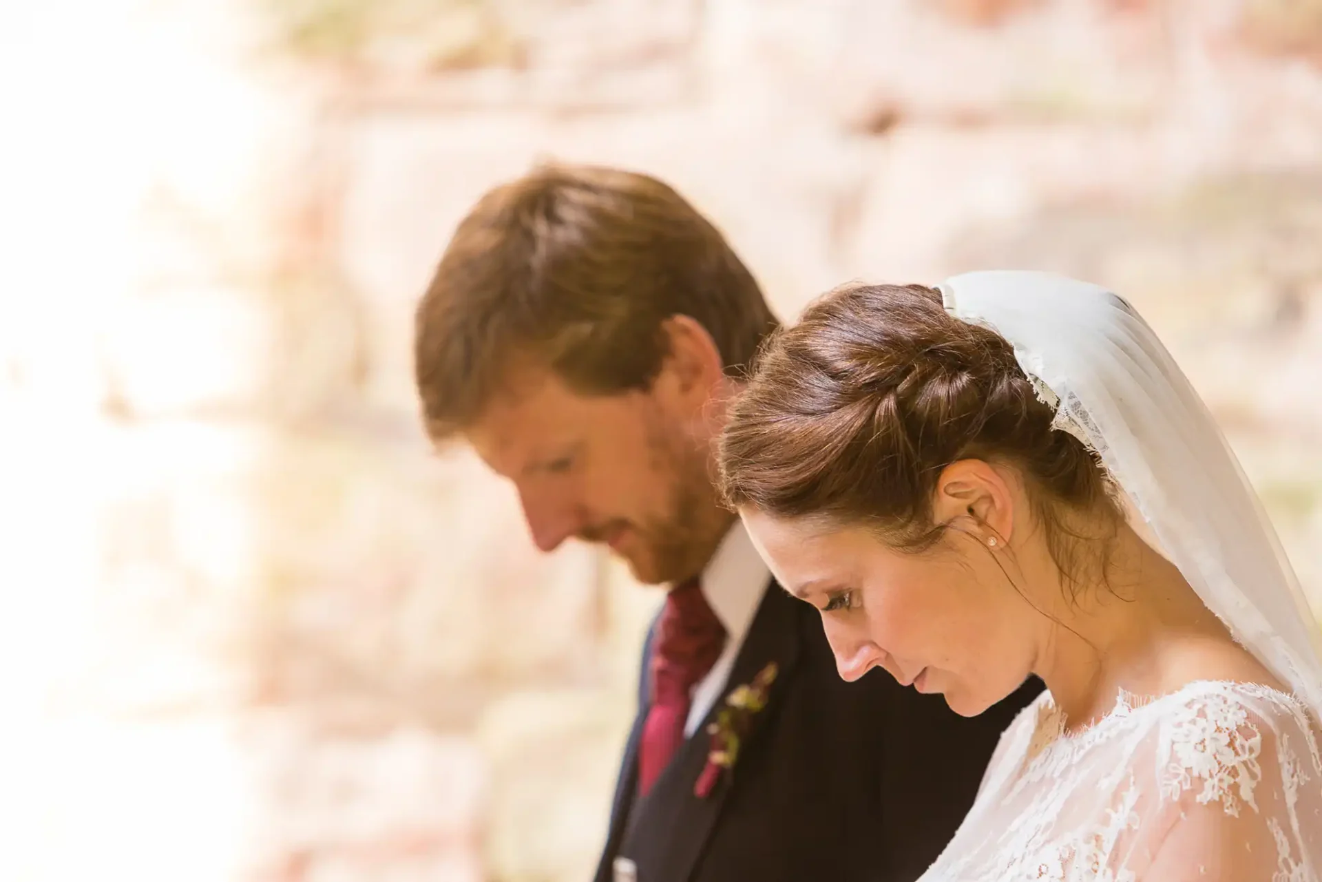 A bride and groom standing together, heads bowed, in soft focus with a warm, light background.