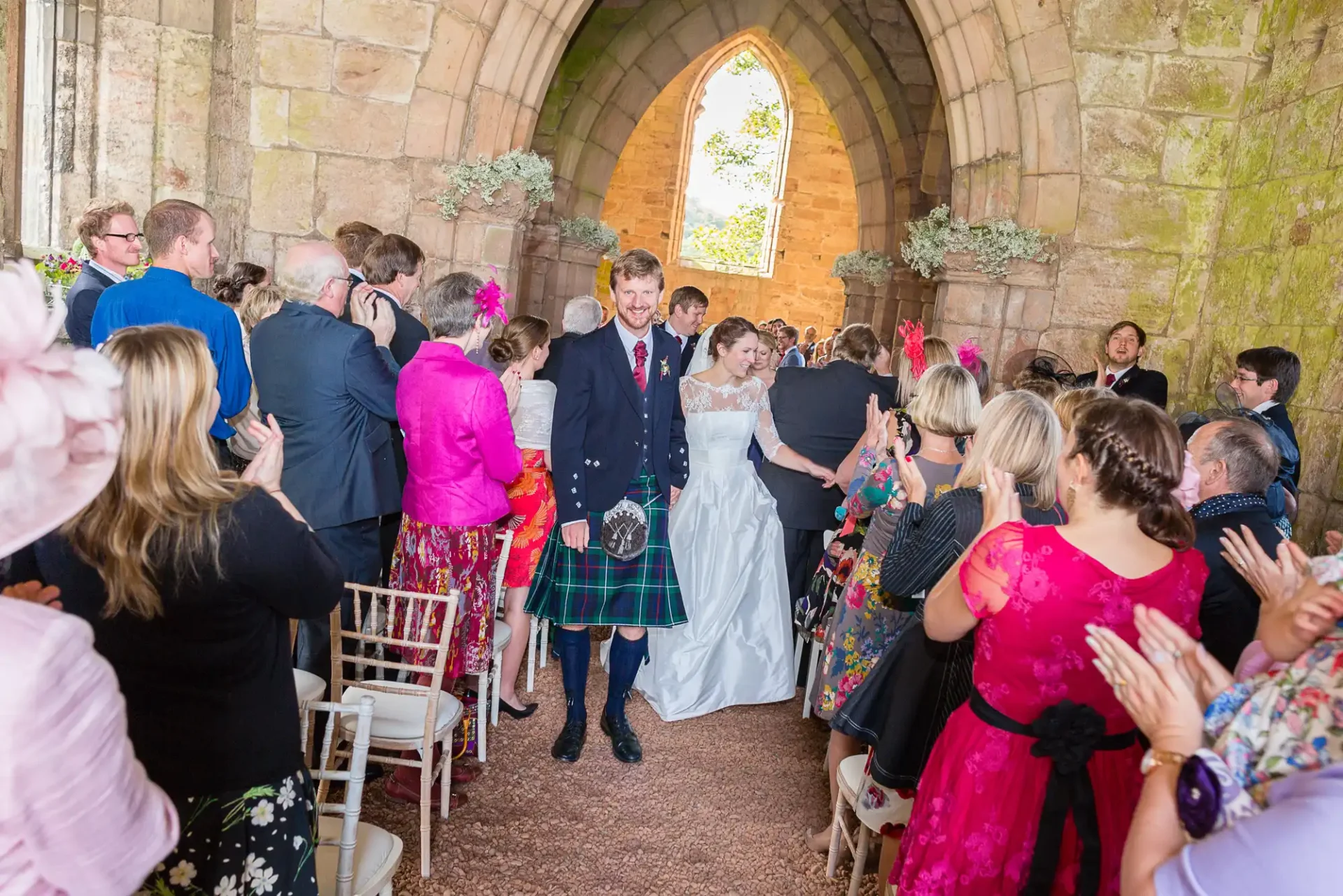 Bride and groom walking down the aisle in a church, surrounded by guests clapping, the groom in a kilt and the bride in a white dress.