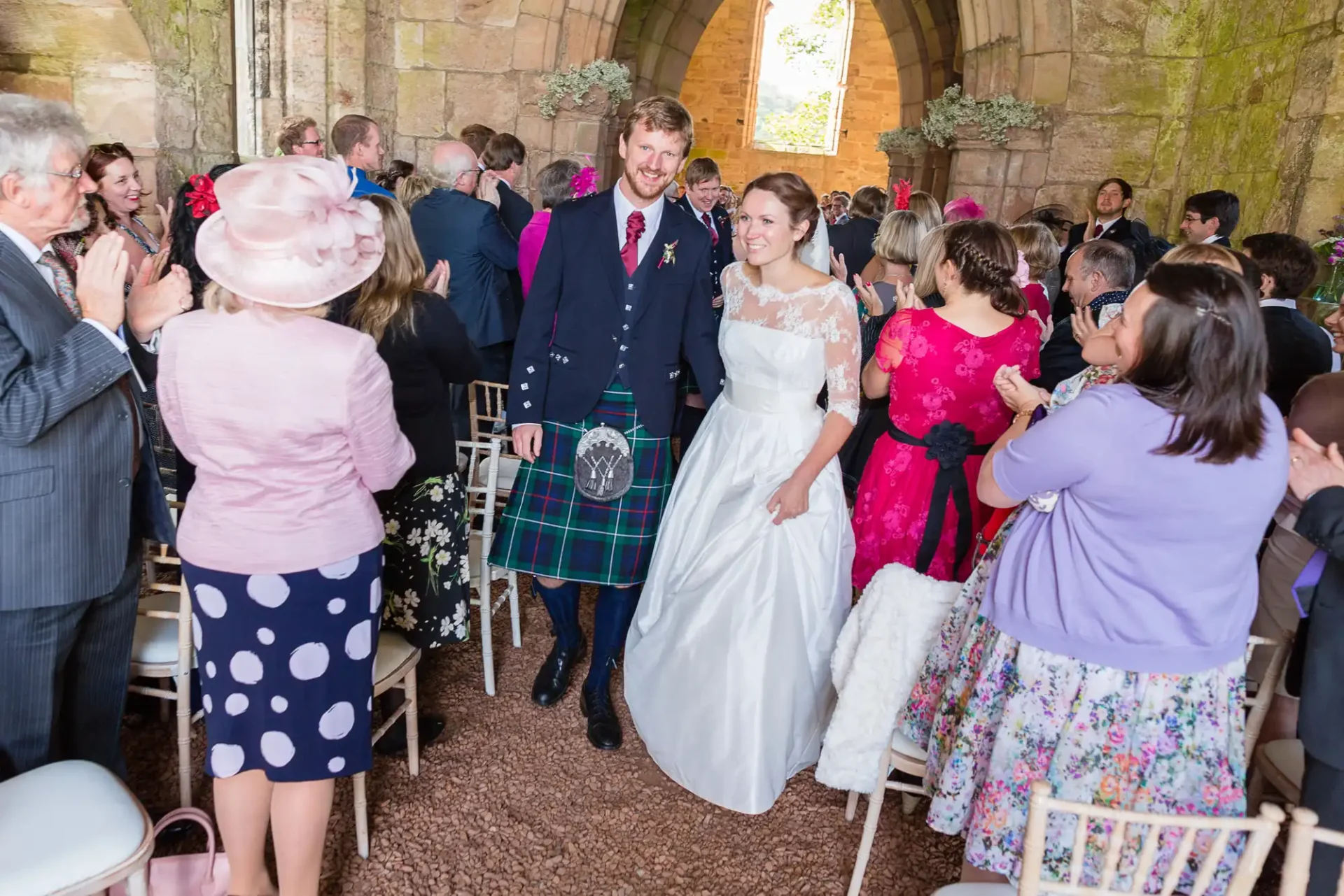 Bride and groom smiling as they walk through guests clapping inside a historic church, the groom wearing a kilt.