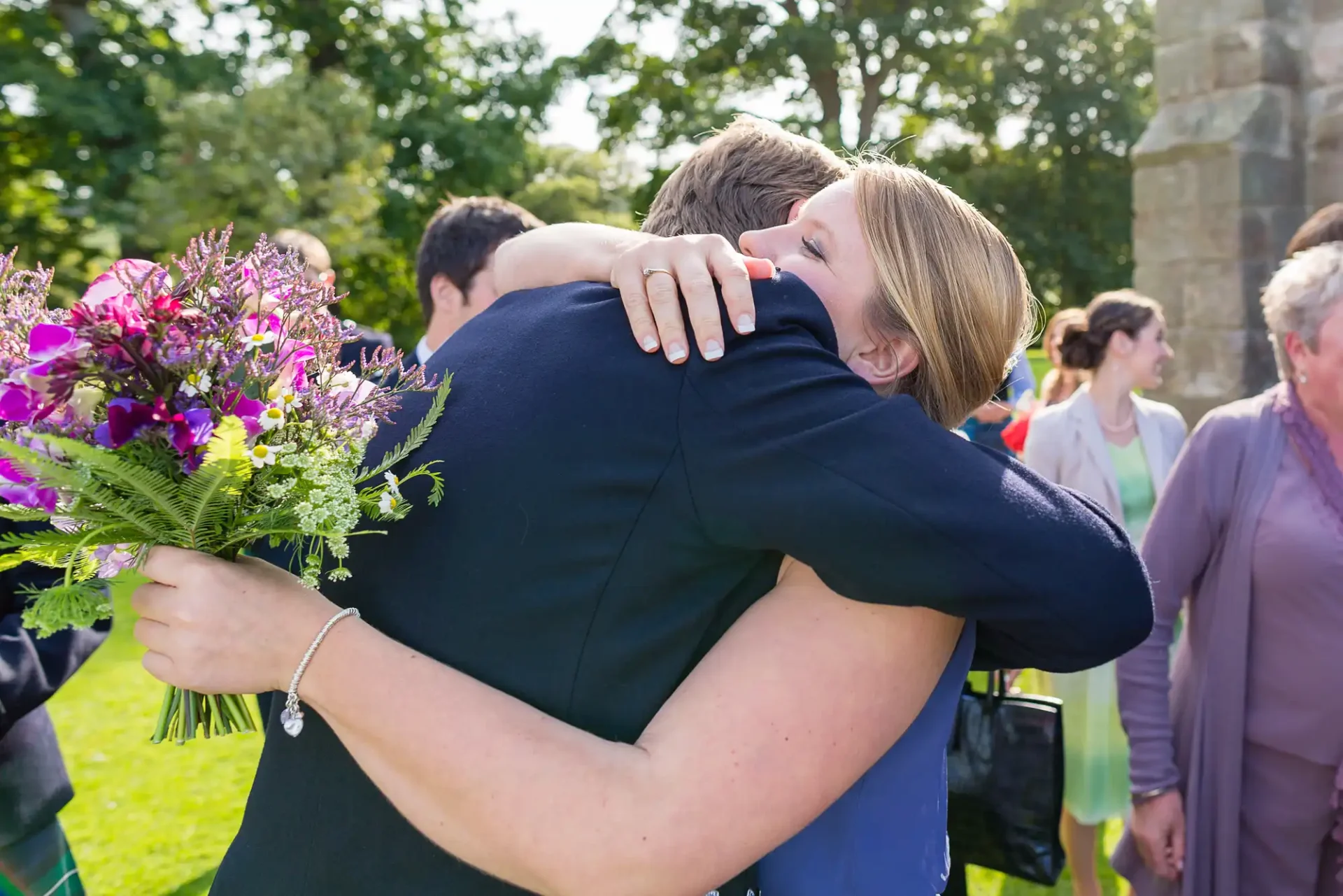A woman clutching a bouquet of purple flowers embraces a man in a navy suit outdoors, surrounded by other people in a sunny park setting.