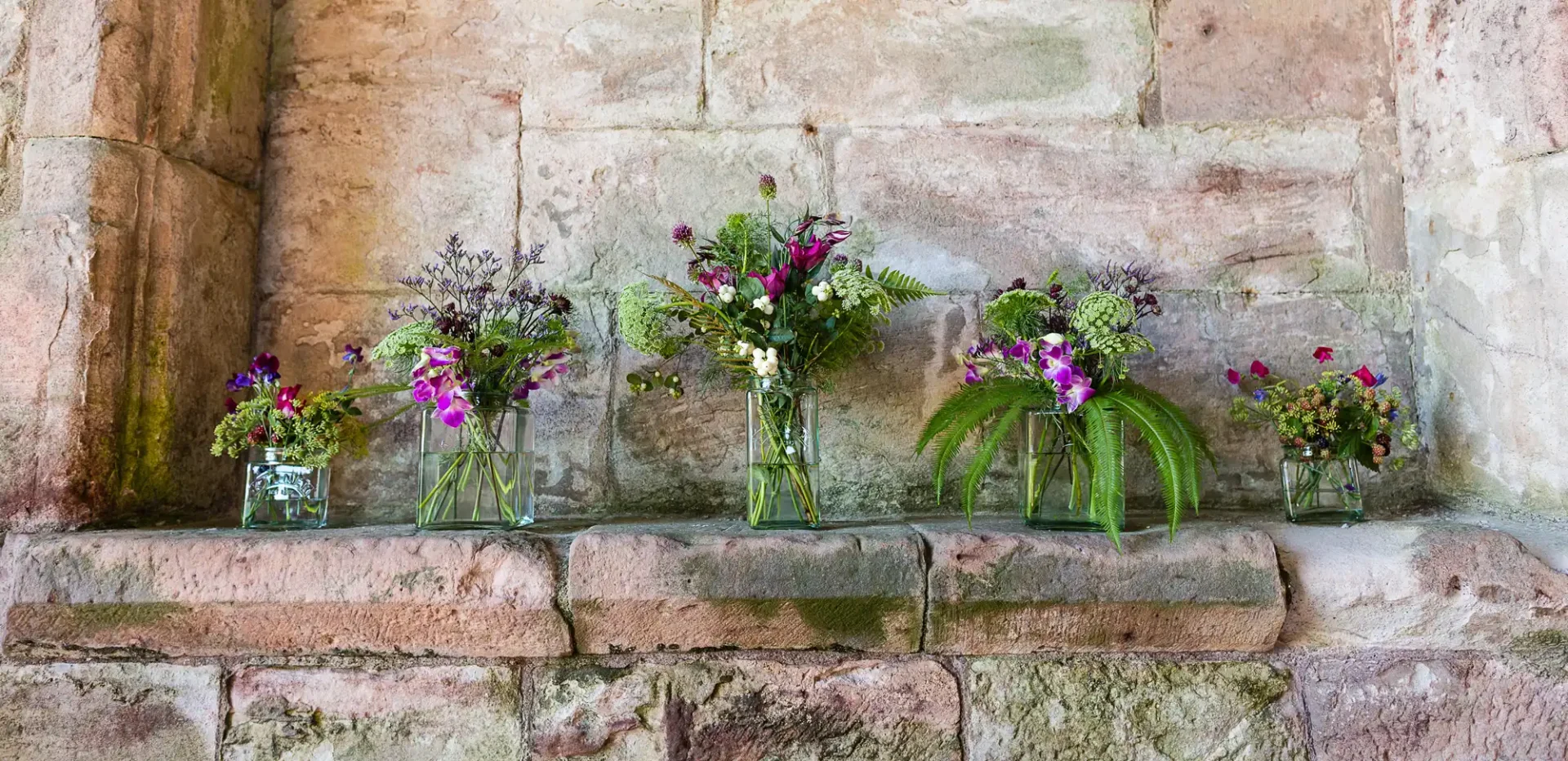 Five assorted flower arrangements in glass vases on a weathered stone ledge.