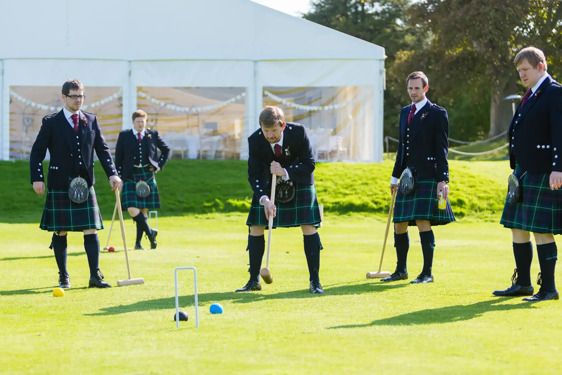Four men in traditional kilts playing croquet on a sunny, grassy field, with one actively taking a shot while the others watch.