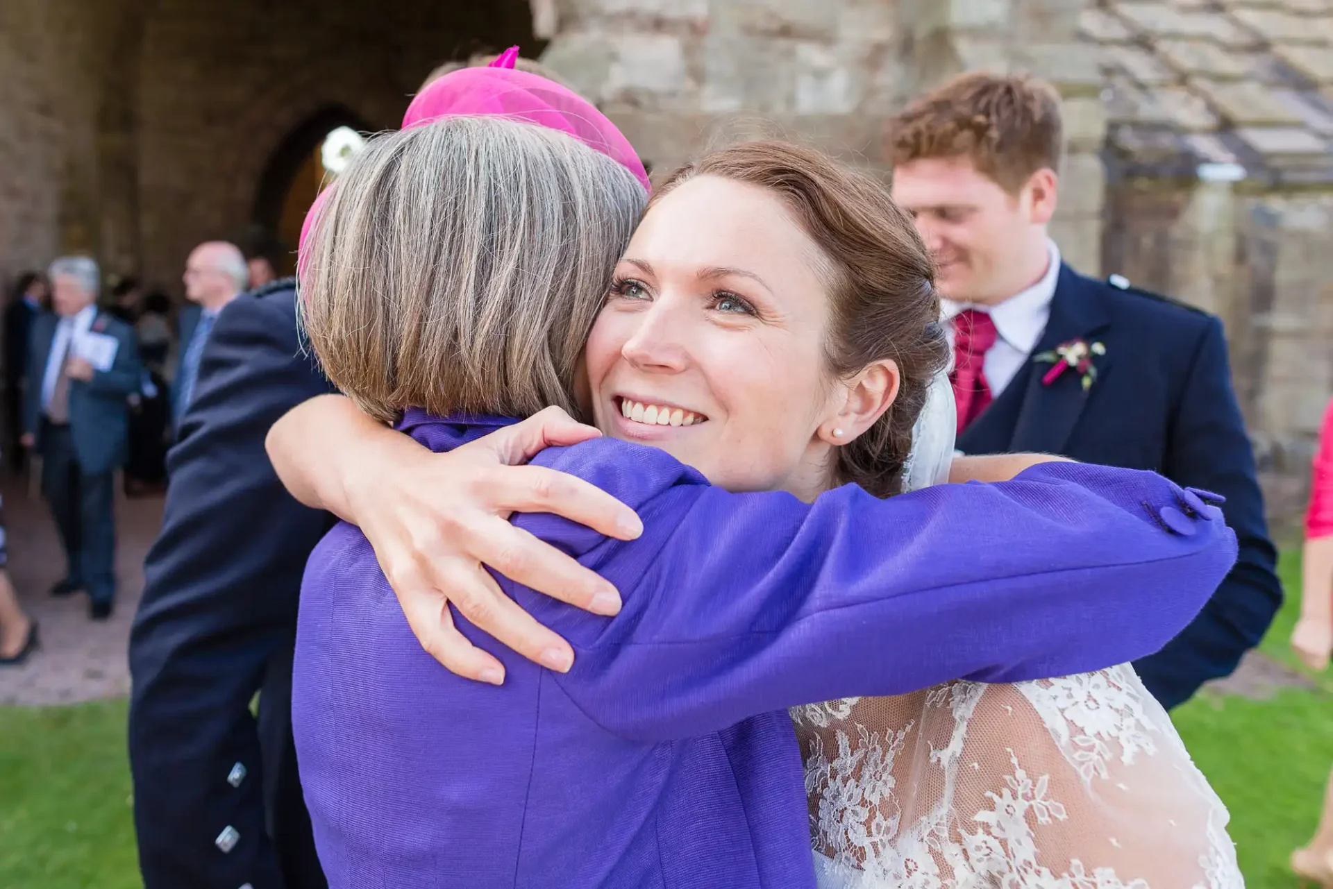 A bride in a lace dress joyfully hugs an older woman in a purple jacket at a wedding, with guests and a stone building in the background.